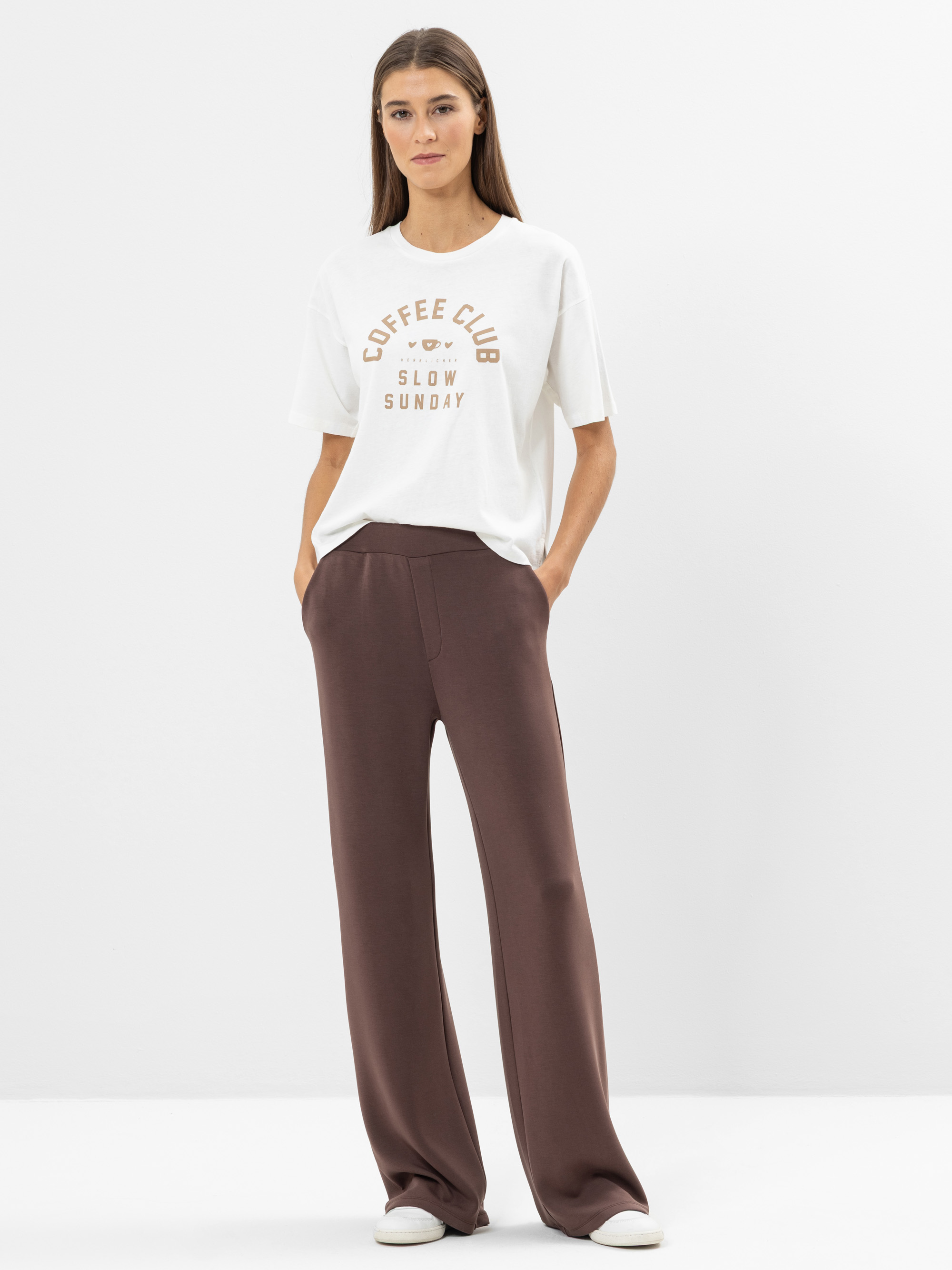 Eine Frau steht vor einem schlichten weißen Hintergrund und trägt ein weißes T-Shirt mit der Aufschrift COFFEE CLUB SLOW SUNDAY, eine weite braune Hose und weiße Turnschuhe. Ihre Hände stecken in den Hosentaschen und sie schaut ruhig in die Kamera.