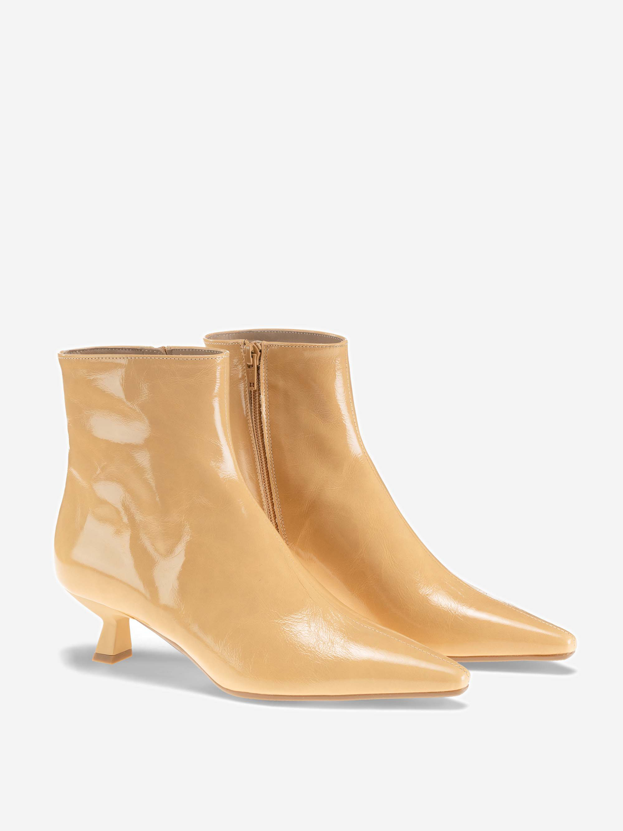 Ein Paar glänzender, hellbeiger Stiefeletten mit spitzer Spitze, niedrigem Kitten-Heel und seitlichen Reißverschlüssen, die vor einem schlichten weißen Hintergrund stehen.