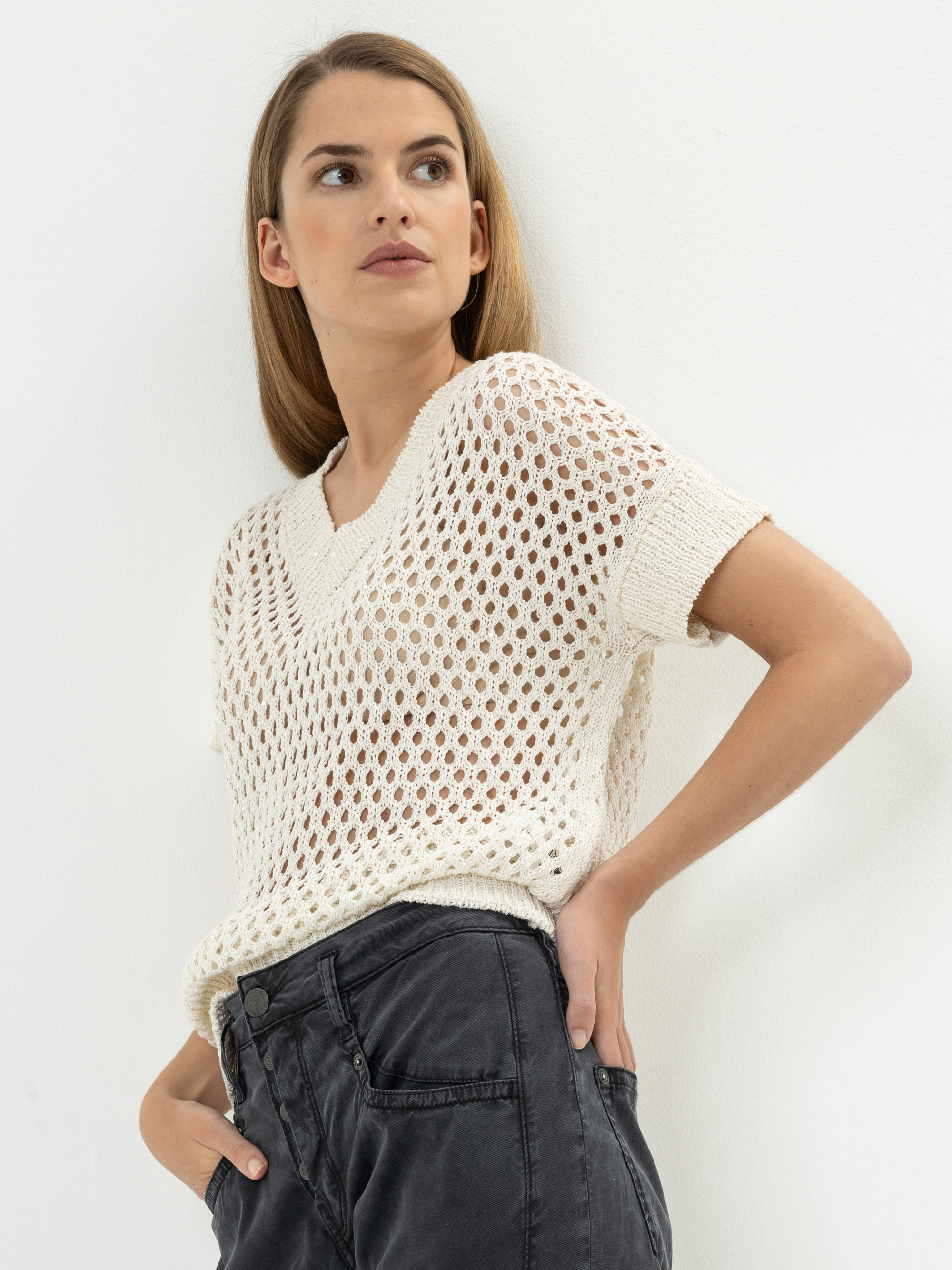 Eine Frau steht vor einer weißen Wand und trägt einen weißen, kurzärmeligen, offen gestrickten Pullover über einem hellen Oberteil und eine dunkle, hoch sitzende Hose. Sie blickt nach links, eine Hand in der Tasche, das Haar offen.