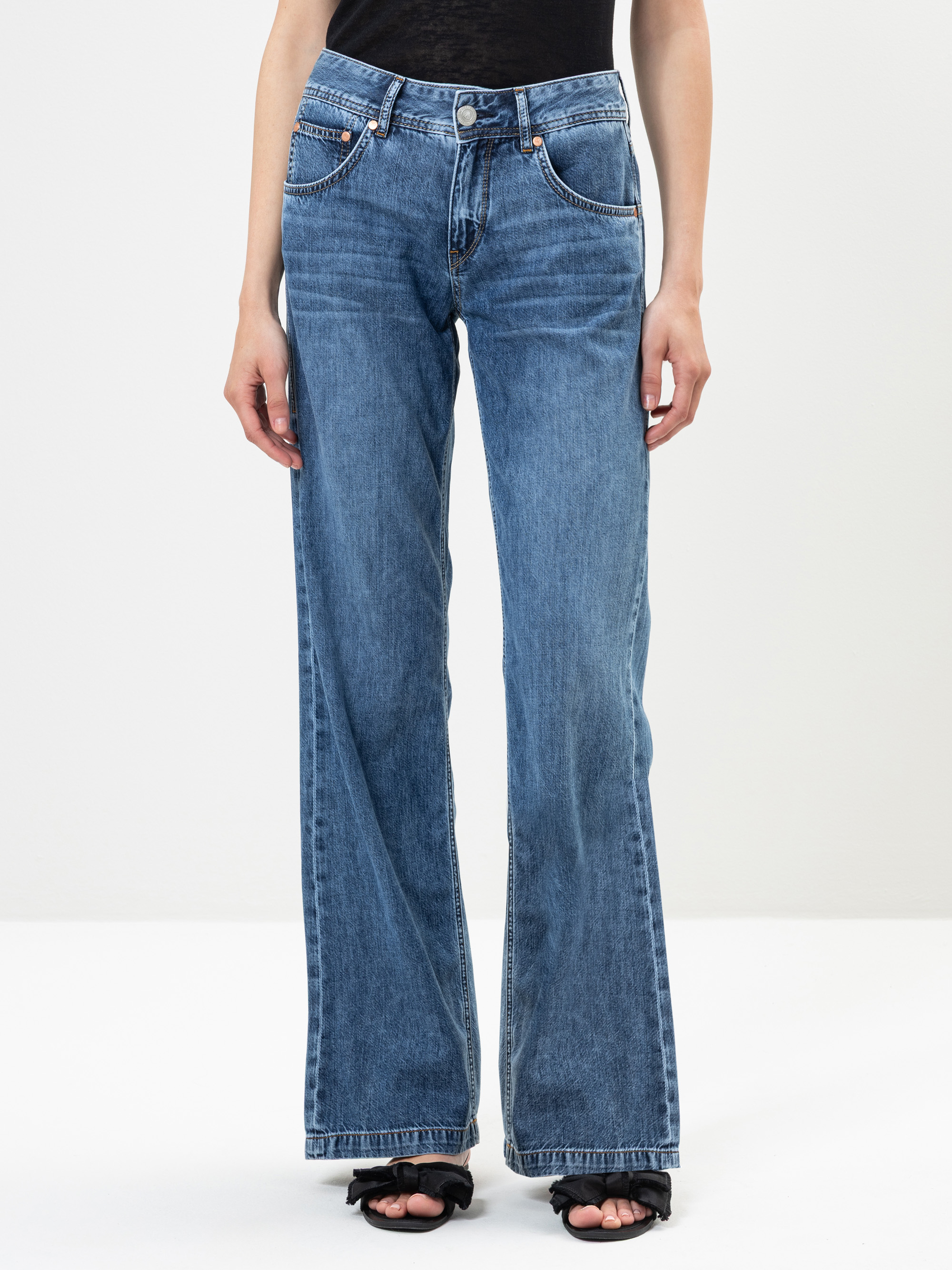 Eine Person, die eine blaue, weit geschnittene Jeans mit Vorder- und Gesäßtaschen trägt, steht vor einem schlichten weißen Hintergrund. Nur der untere Teil des Oberkörpers und die Beine sind sichtbar, und die Person trägt schwarze Sandalen mit offen