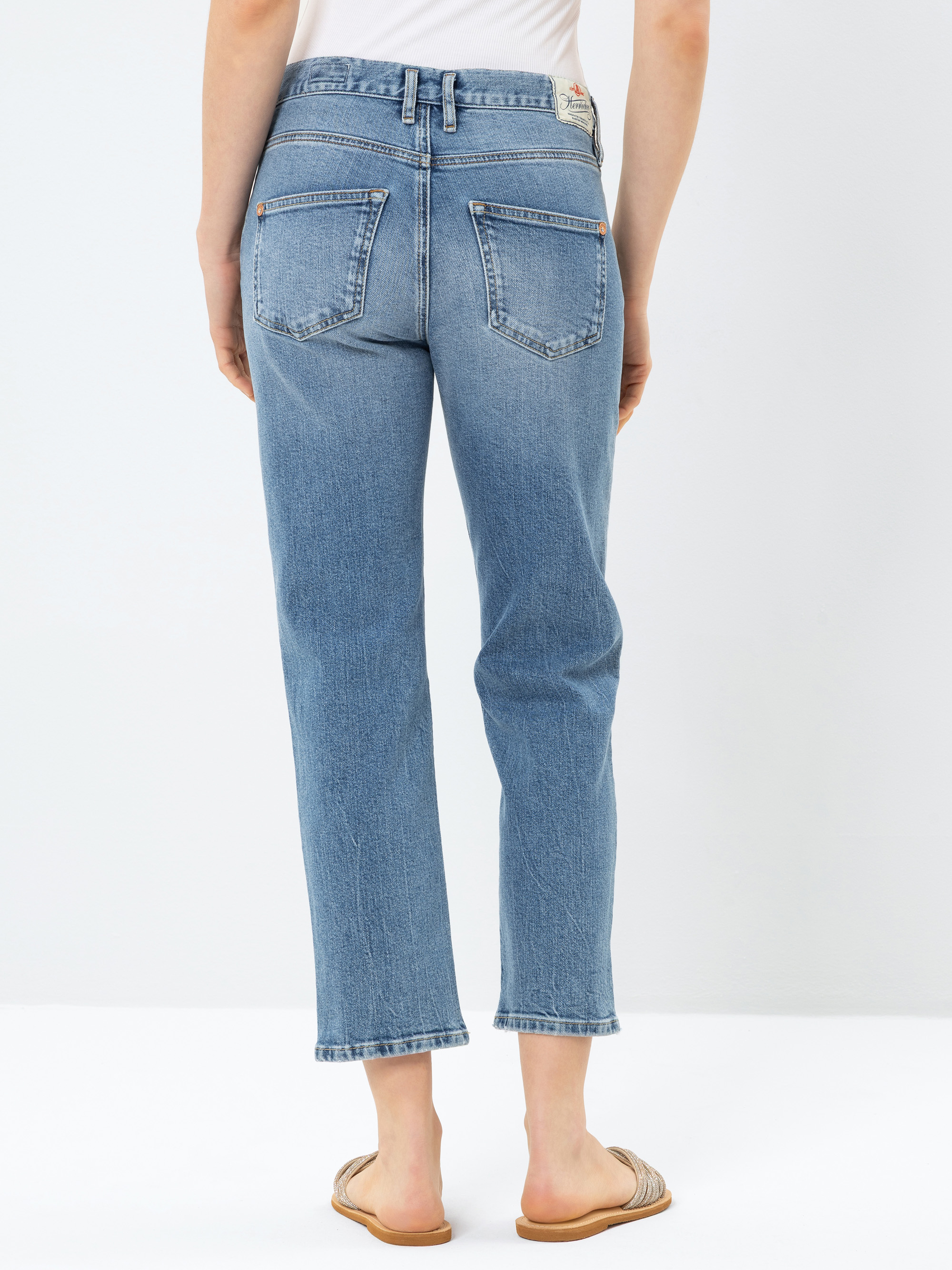 Eine Person steht mit dem Gesicht zur Wand und trägt eine hellblaue Jeans mit hoher Taille und geradem Bein sowie hellbraune flache Sandalen. Die Jeans hat Gesäßtaschen und einen Aufnäher am Bund. Der Hintergrund ist einfarbig weiß.
