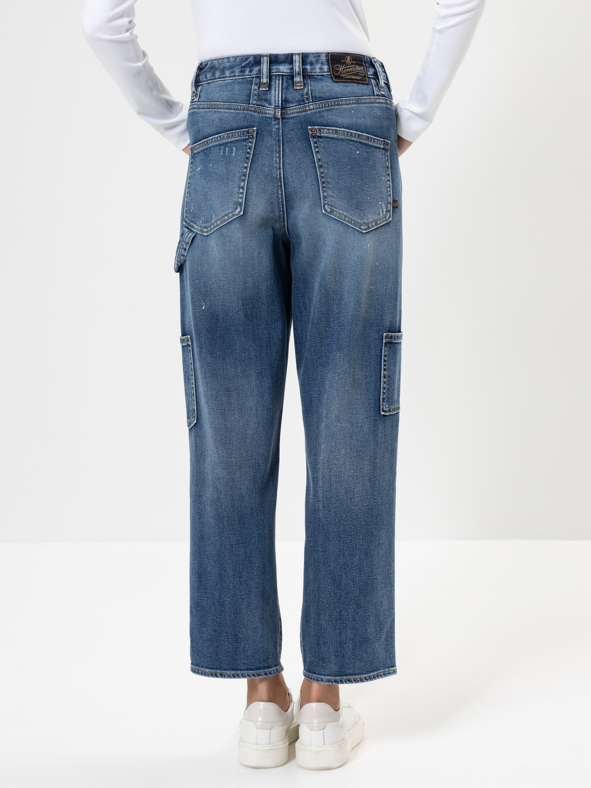 Eine Person, die ein weißes langärmeliges Hemd, eine blaue Jeans mit geradem Bein und großen Gesäßtaschen sowie weiße Turnschuhe trägt, steht mit dem Rücken zur Kamera vor einem schlichten weißen Hintergrund.