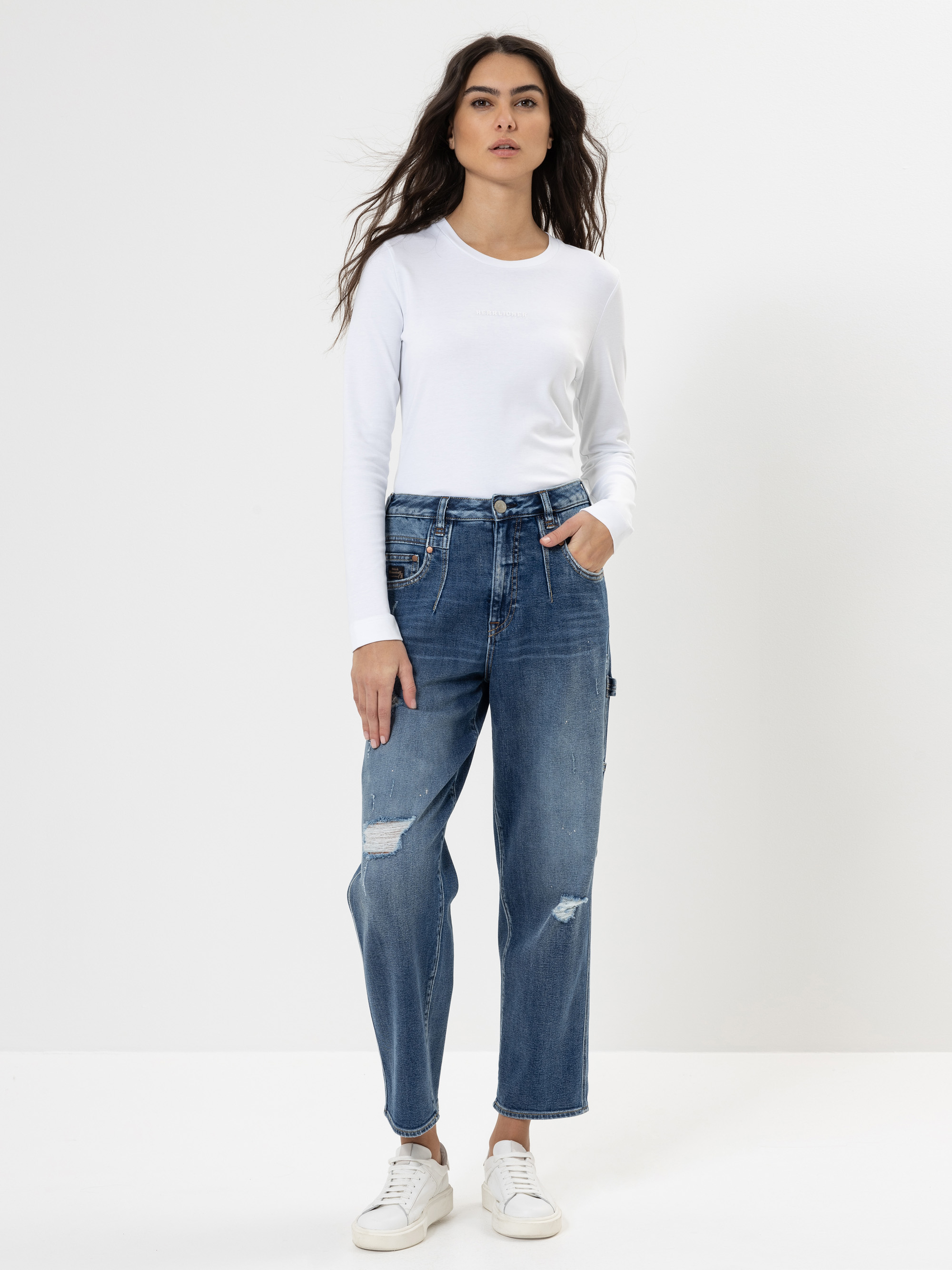 Eine Frau steht vor einem einfarbig weißen Hintergrund und trägt ein weißes, langärmeliges Hemd, eine hochtaillierte Bluejeans mit Rissen und weiße Turnschuhe. Sie hat langes, gewelltes Haar und eine Hand in der Tasche.