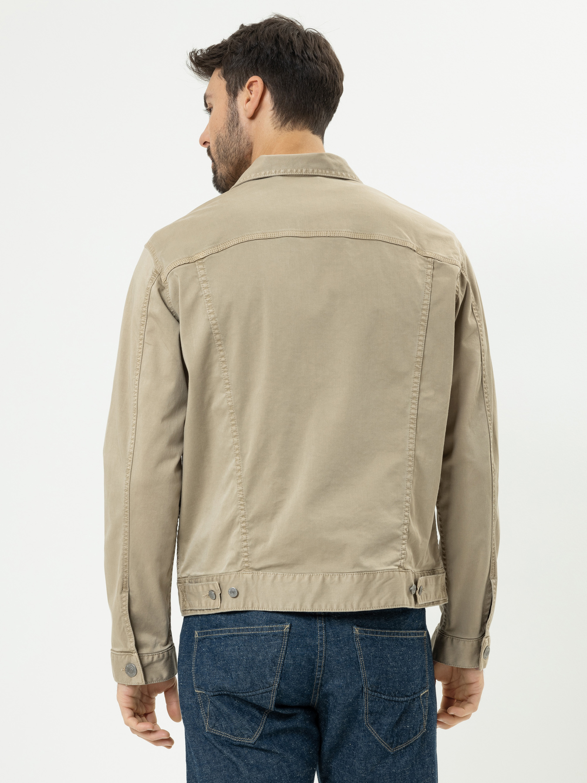 Ein Mann, der eine hellbeige Jeansjacke und blaue Jeans trägt, steht mit dem Gesicht von der Kamera weg vor einem schlichten weißen Hintergrund.