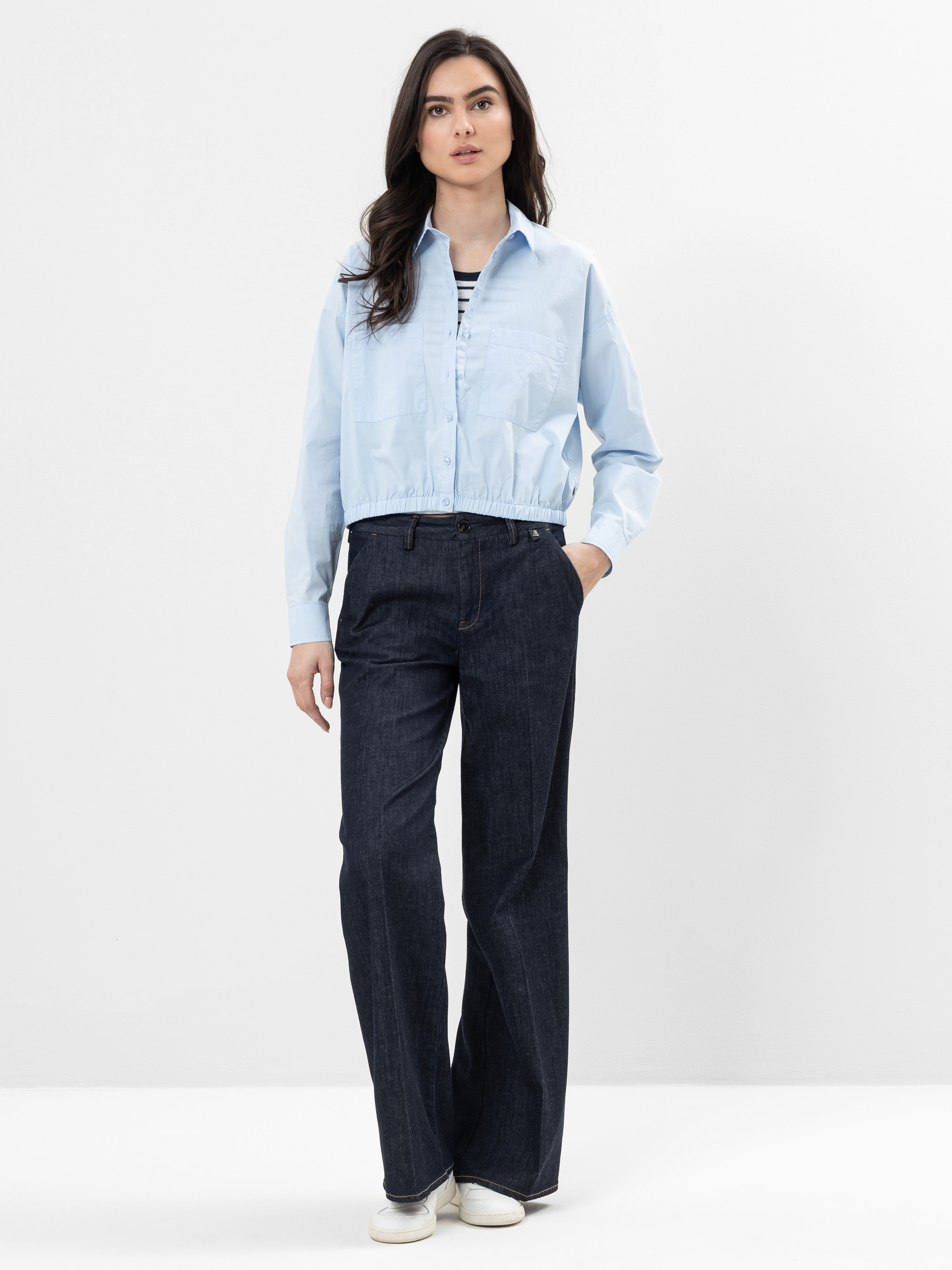 Eine Frau steht vor einem schlichten weißen Hintergrund und trägt eine hellblaue Kurzjacke über einem gestreiften Hemd, dunkle Jeans mit weitem Bein und weiße Schuhe. Sie hat langes dunkles Haar und einen neutralen Gesichtsausdruck.