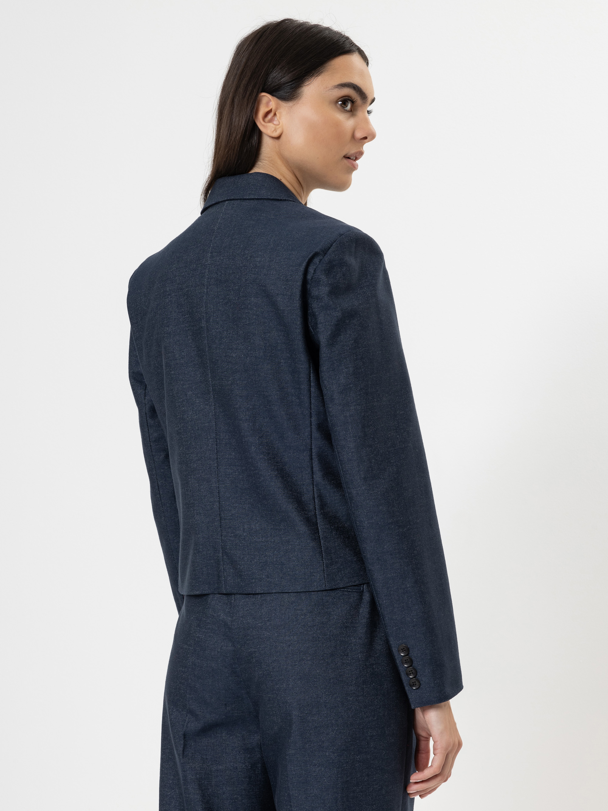 Eine Frau mit langen dunklen Haaren, die eine marineblaue Anzugsjacke und eine dazu passende Hose trägt, steht mit leicht zur Seite gedrehtem Kopf vor einem schlichten weißen Hintergrund.
