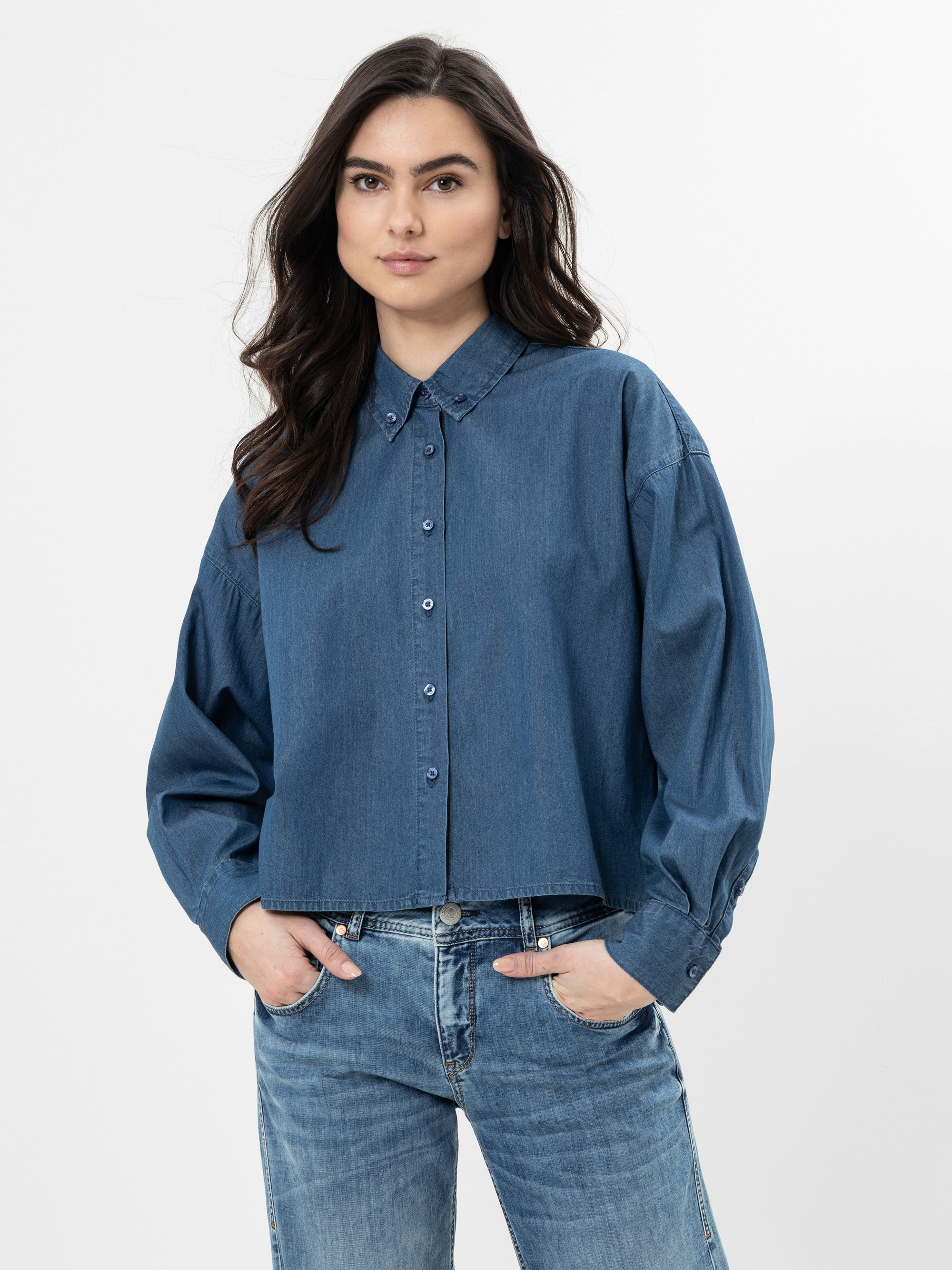 Eine Frau mit langen dunklen Haaren, die ein blaues Jeanshemd und blaue Jeans trägt, steht mit den Händen in den Taschen vor einem schlichten weißen Hintergrund.