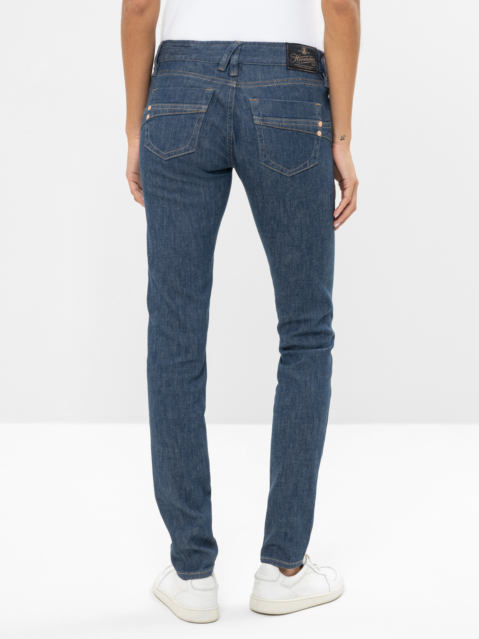Eine Person in blauen Skinny-Jeans und weißen Turnschuhen ist von hinten zu sehen und steht vor einem schlichten weißen Hintergrund.