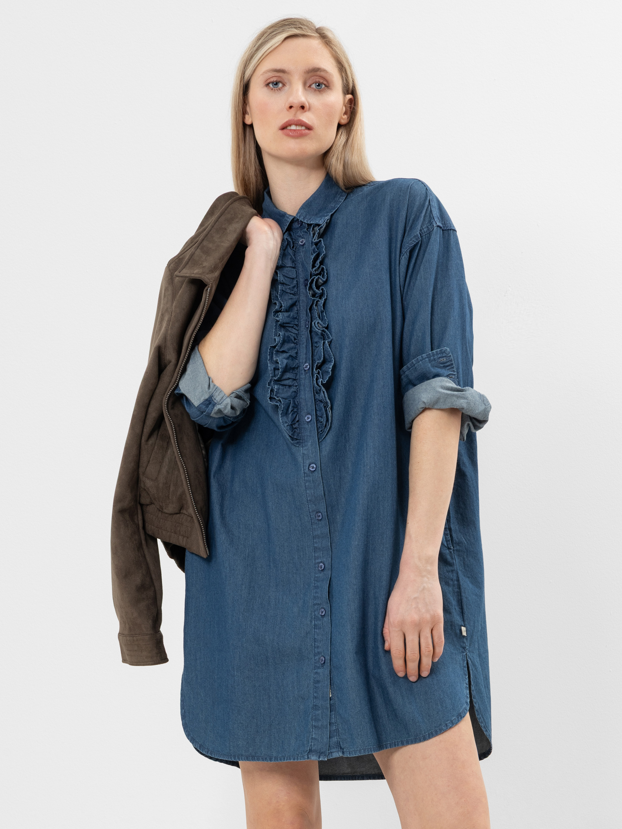 Eine Frau steht vor einem schlichten Hintergrund und trägt ein blaues Hemdkleid aus Jeansstoff mit Rüschen auf der Vorderseite. Sie trägt eine Jacke über der Schulter, hat die Ärmel hochgekrempelt und blickt selbstbewusst in die Kamera.