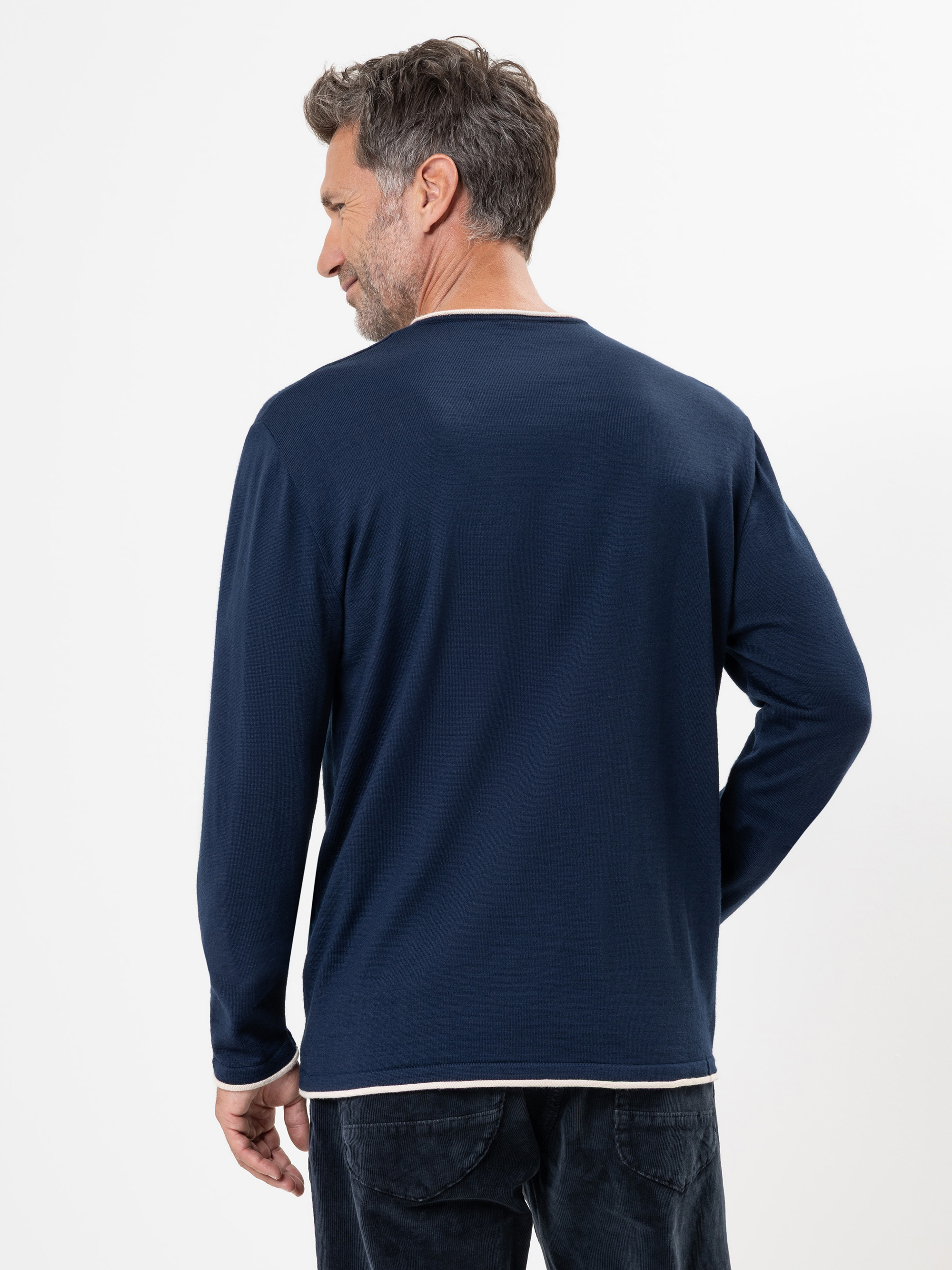 Ein Mann mit kurzen braunen Haaren, der ein langärmeliges marineblaues Hemd und eine dunkle Hose trägt, steht mit dem Gesicht zur Kamera vor einem schlichten weißen Hintergrund.