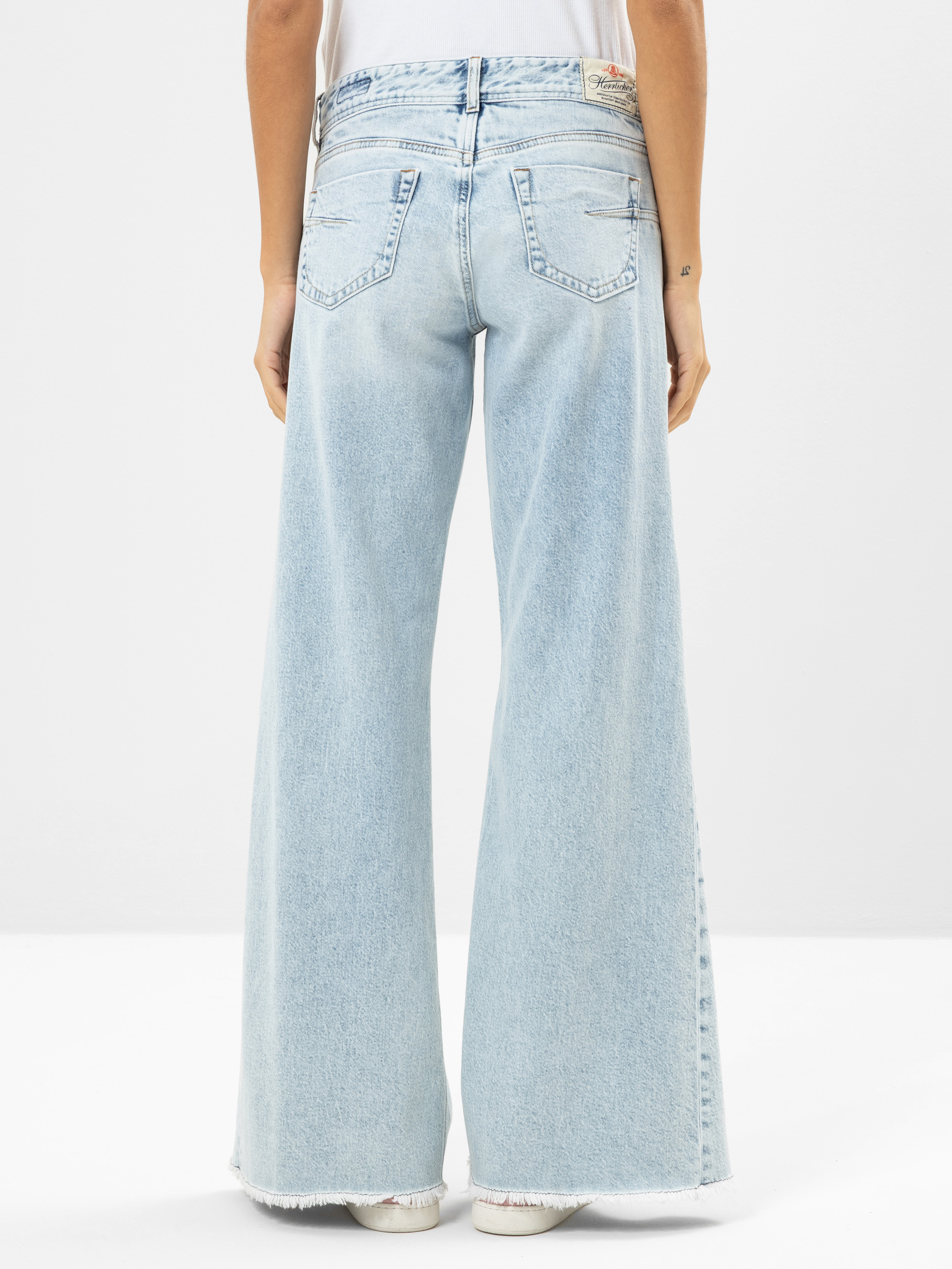Eine Person trägt eine hellblaue, hochtaillierte Jeans mit weitem Bein und ausgefransten Säumen, von hinten gesehen. Die Jeans hat zwei Gesäßtaschen und wird mit einem weißen Oberteil und weißen Schuhen kombiniert.