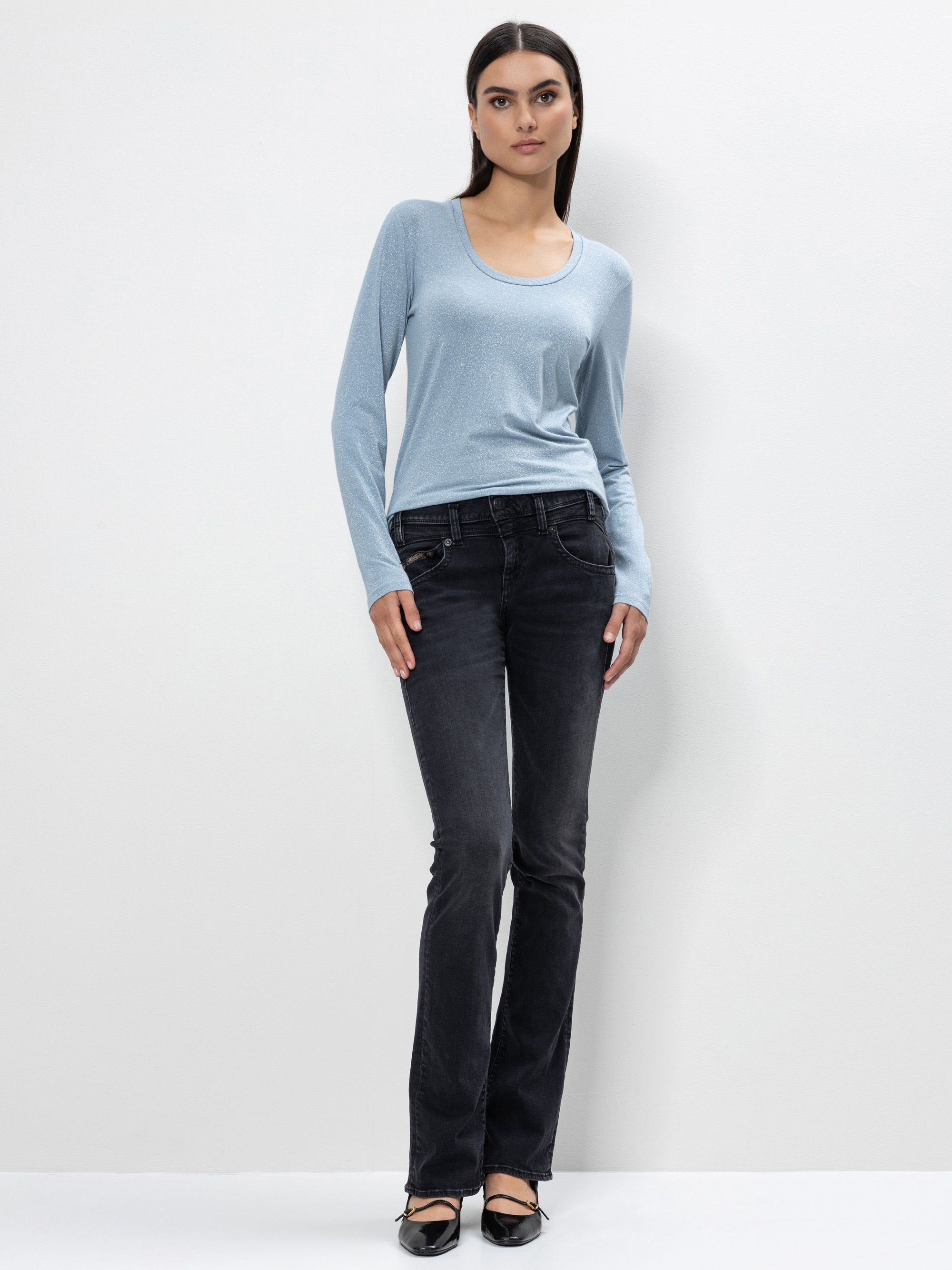 Eine Frau steht vor einem schlichten weißen Hintergrund und trägt ein hellblaues langärmeliges Oberteil, dunkle Jeans mit geradem Bein und schwarze flache Schuhe. Sie hat glattes dunkles Haar und einen neutralen Gesichtsausdruck.