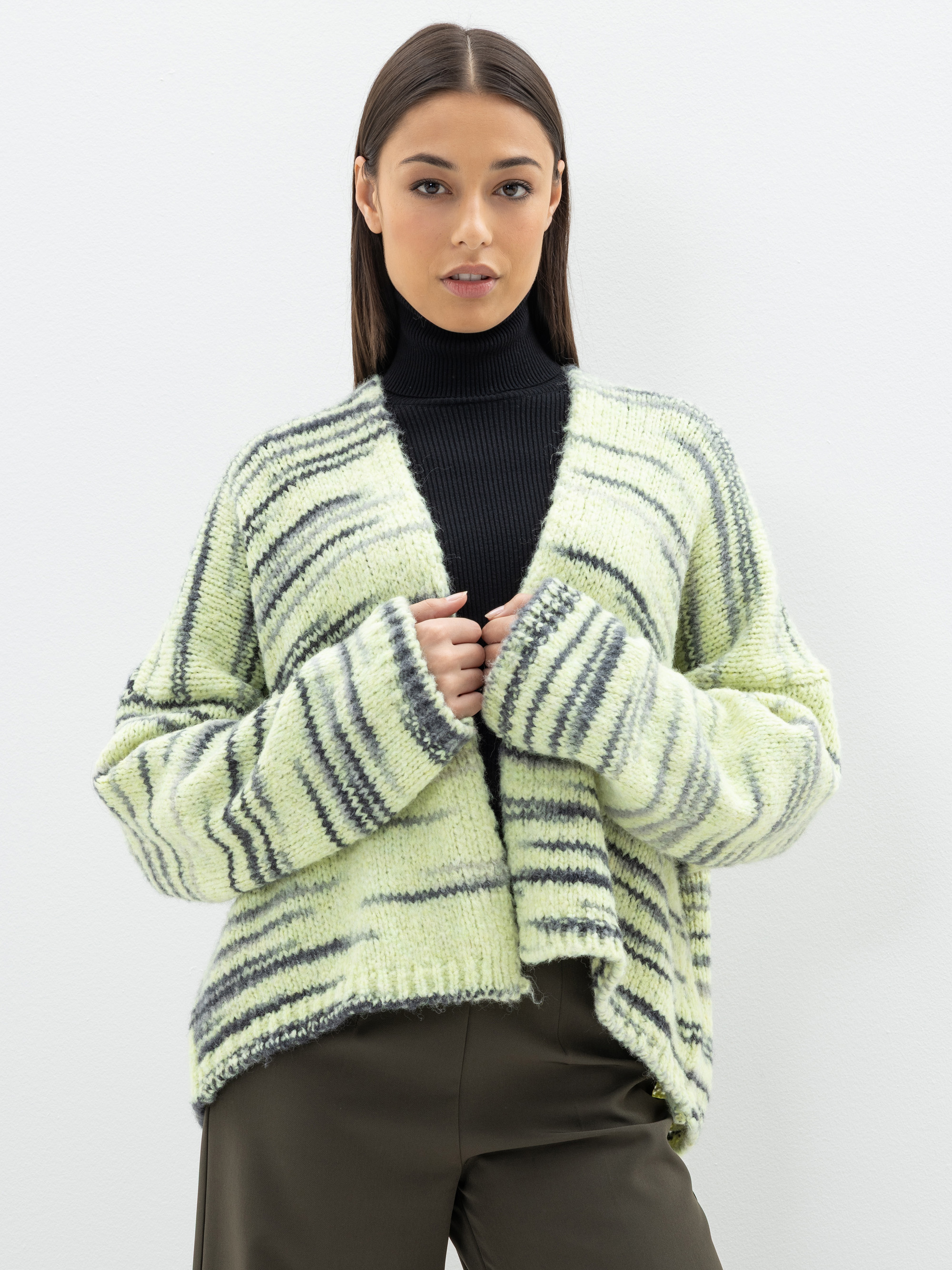Eine Frau mit glattem braunem Haar trägt einen schwarzen Rollkragenpullover, eine übergroße hellgrün-grau gestreifte Strickjacke und eine dunkle Hose und steht vor einem schlichten weißen Hintergrund.