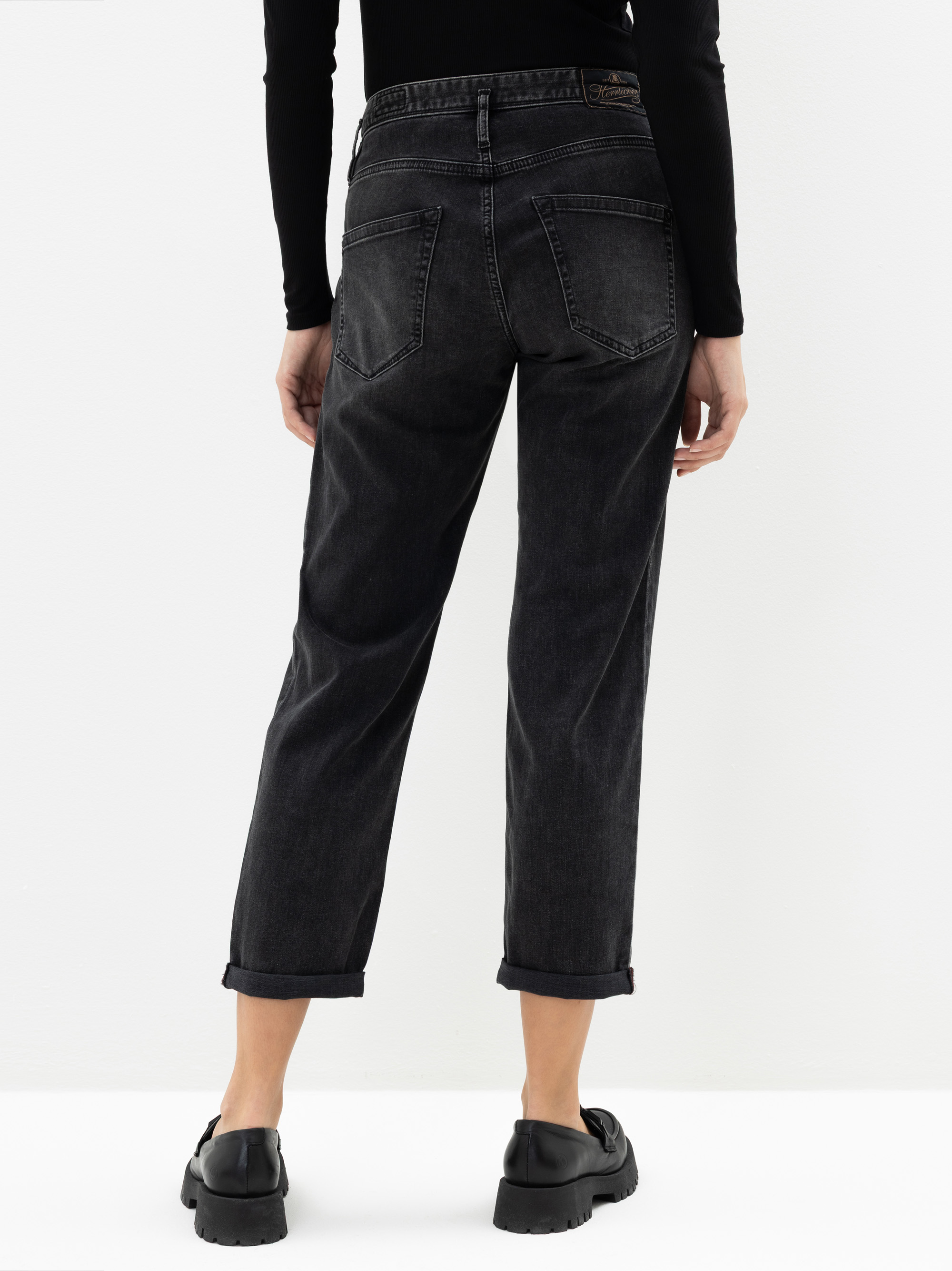 Eine Person, die eine schwarze Jeans mit Bündchen, ein schwarzes langärmeliges Oberteil und schwarze Schuhe trägt, steht mit dem Rücken zur Kamera vor einem weißen Hintergrund.