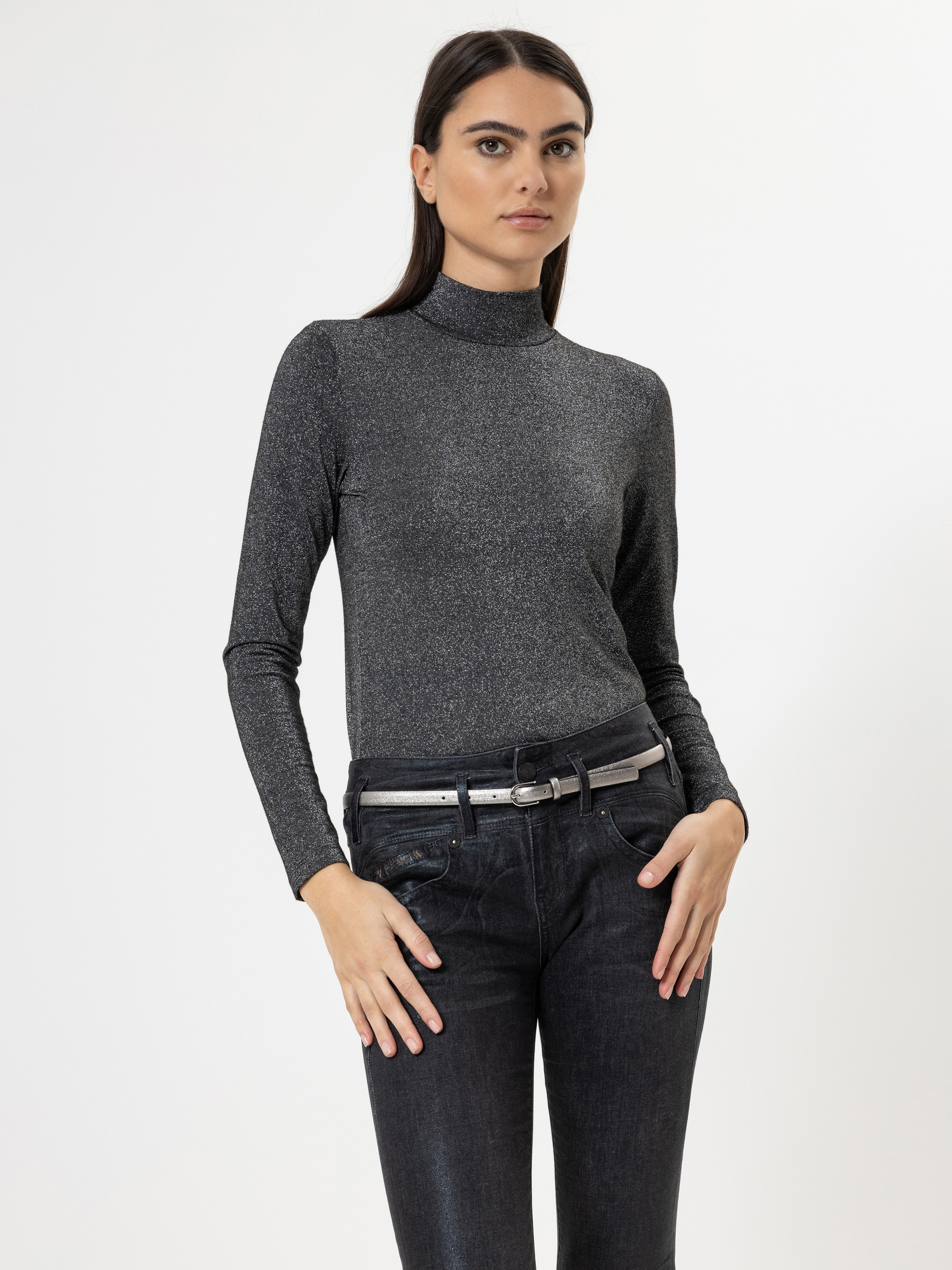Eine Frau mit langen dunklen Haaren trägt ein tailliertes, grau schimmerndes Rollkragenoberteil und eine schwarze Jeans mit einem grauen Gürtel, die vor einem schlichten weißen Hintergrund steht.