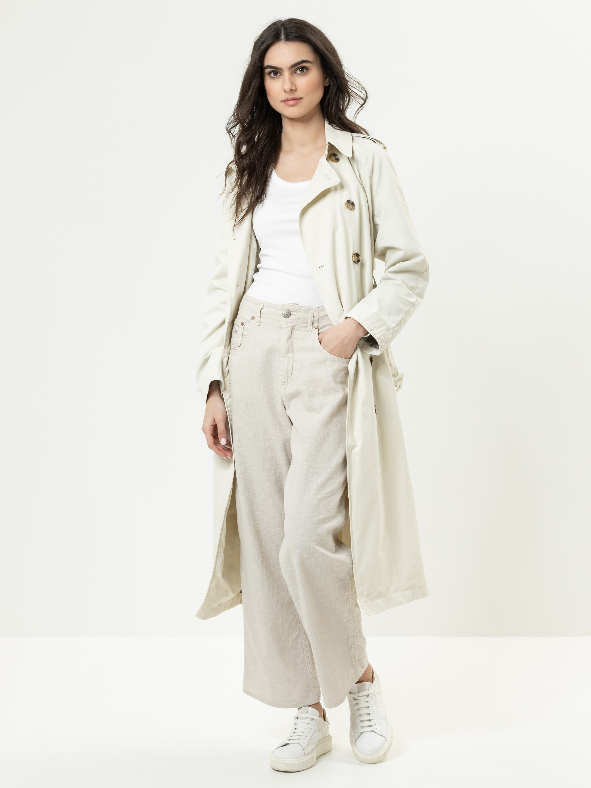 Eine Frau steht vor einem weißen Hintergrund und trägt einen langen, hellbeigen Trenchcoat über einem weißen Oberteil und eine beigefarbene Hose mit weitem Bein, dazu weiße Turnschuhe. Sie hat langes dunkles Haar und eine Hand in der Tasche.