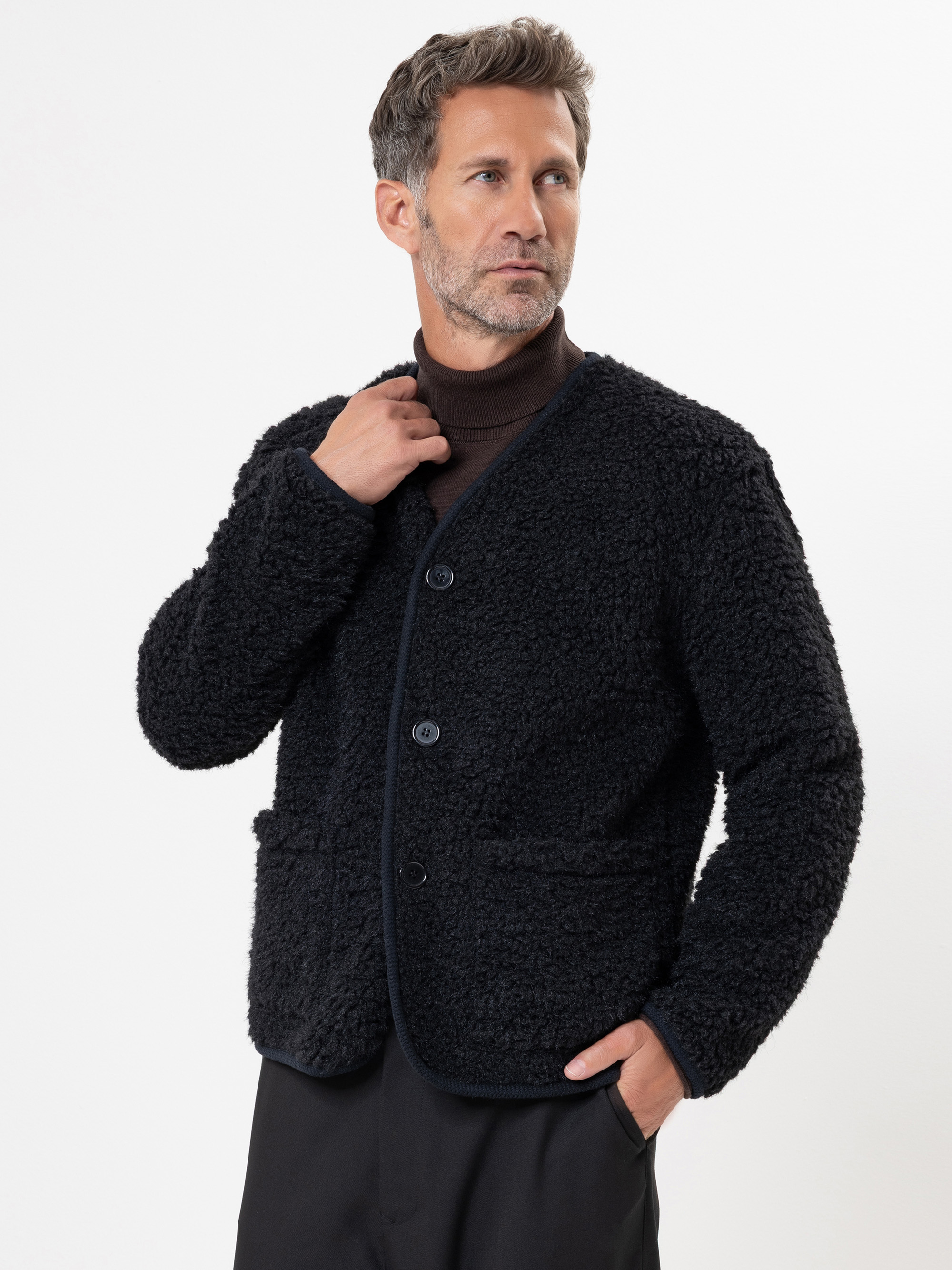 Ein Mann mit kurzen, gewellten grauen Haaren und einem Bart trägt eine strukturierte schwarze Jacke mit Knöpfen über einem dunklen Rollkragenpullover. Er steht leicht nach links gewandt, mit einer Hand am Kragen, vor einem schlichten weißen Hintergrun