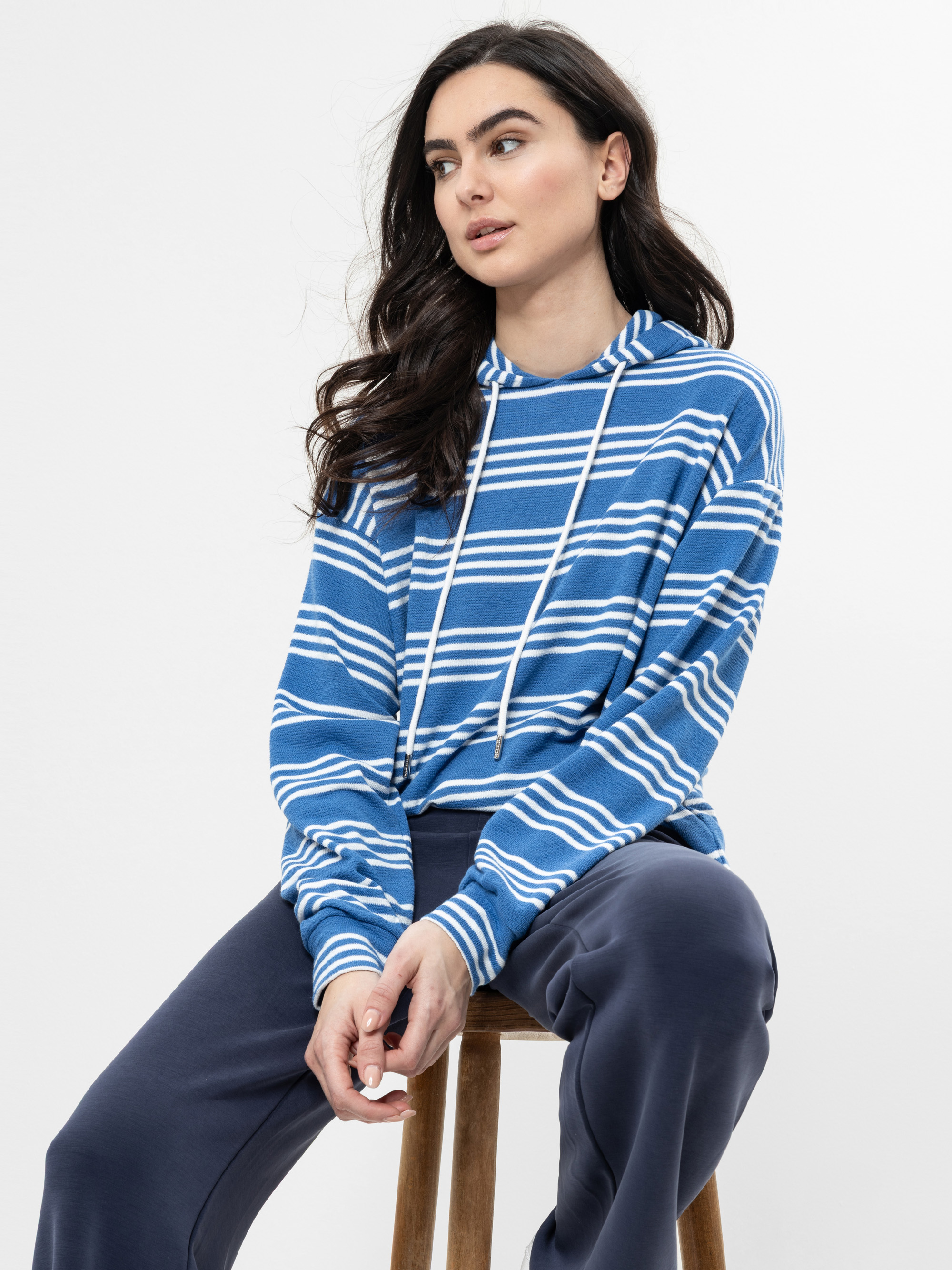 Eine Frau mit langen dunklen Haaren, die einen blau-weiß gestreiften Kapuzenpulli und eine marineblaue Hose trägt, sitzt auf einem Holzhocker vor einem schlichten weißen Hintergrund und schaut leicht zur Seite.