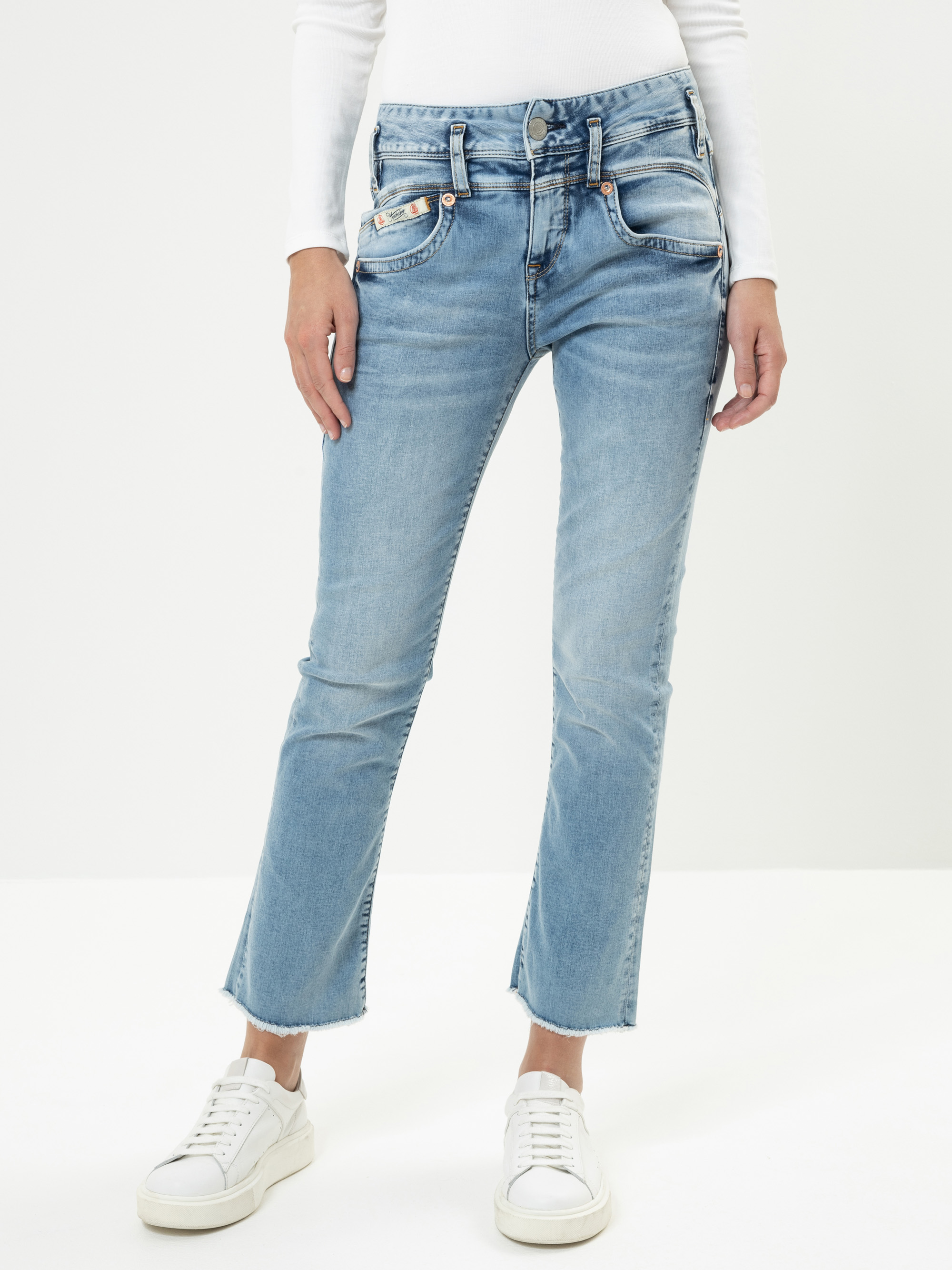 Eine Person, die hellblaue abgeschnittene Jeans, ein weißes langärmeliges Oberteil und weiße Turnschuhe trägt, steht vor einem schlichten weißen Hintergrund. Das Foto ist an den Schultern abgeschnitten.