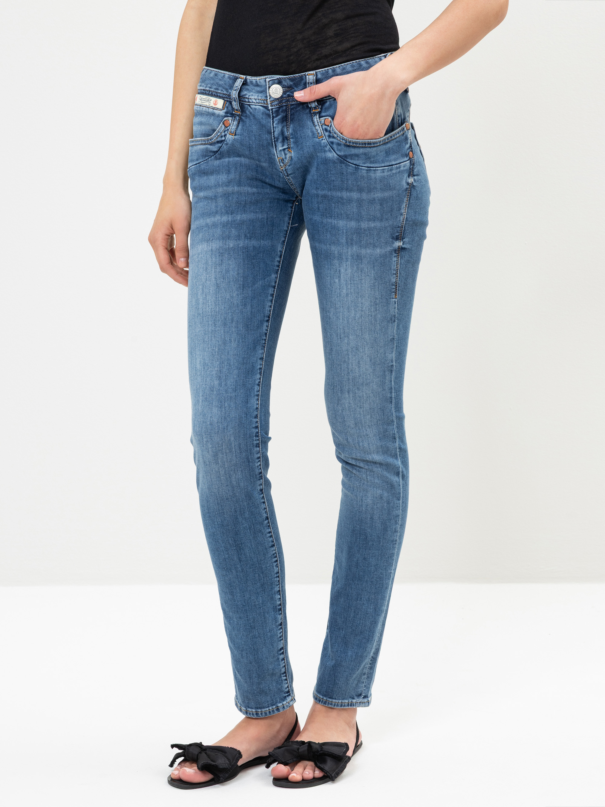 Eine Person, die hellblaue Röhrenjeans, ein schwarzes Oberteil und schwarze, flache, offene Schuhe mit Schleifen trägt, steht vor einem schlichten, hellen Hintergrund. Nur der Körper von den Schultern bis zu den Füßen ist sichtbar.