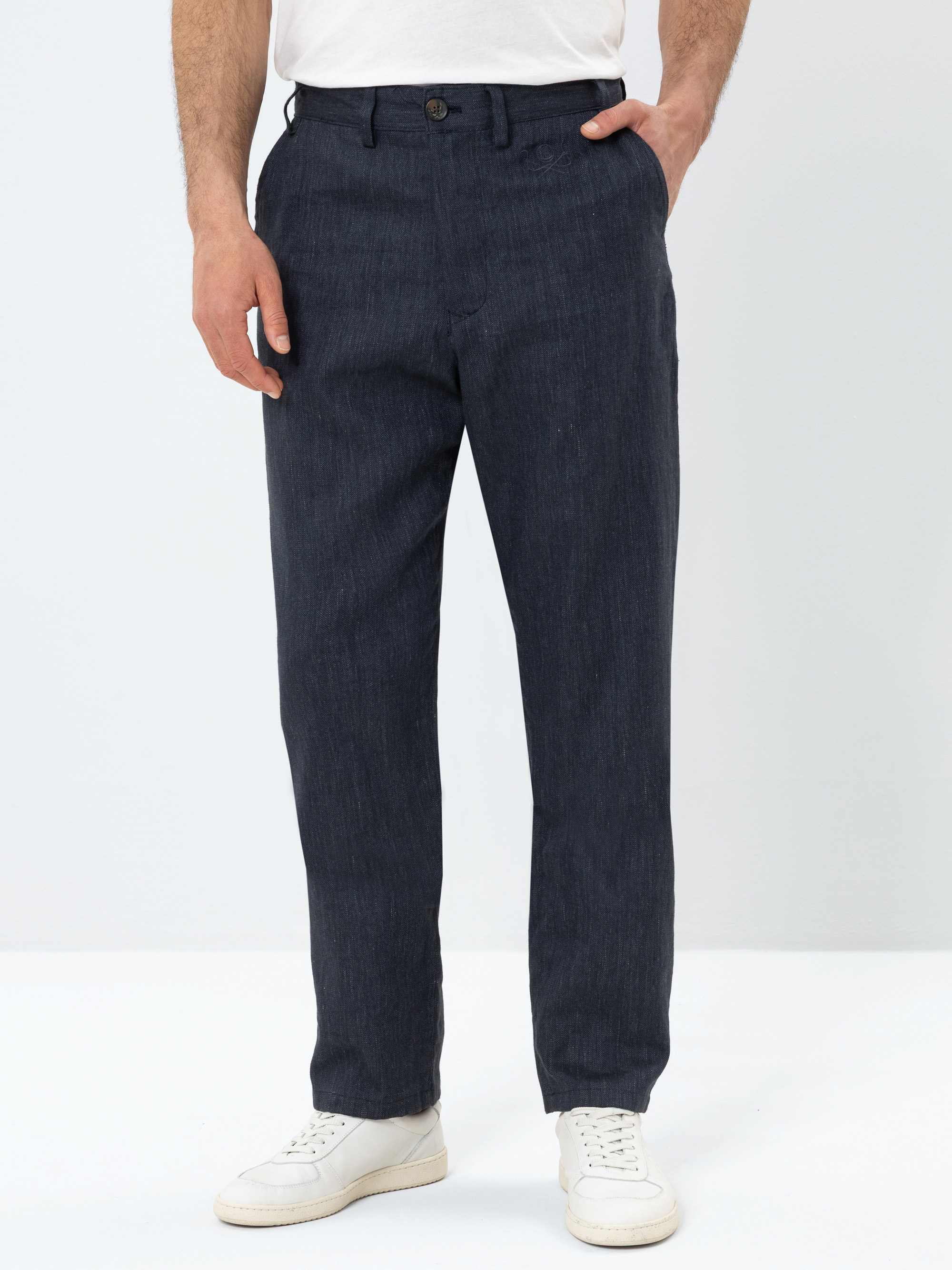 Eine Person, die eine dunkelblaue Hose, ein weißes T-Shirt und weiße Turnschuhe trägt, steht mit einer Hand in der Tasche vor einem schlichten weißen Hintergrund.