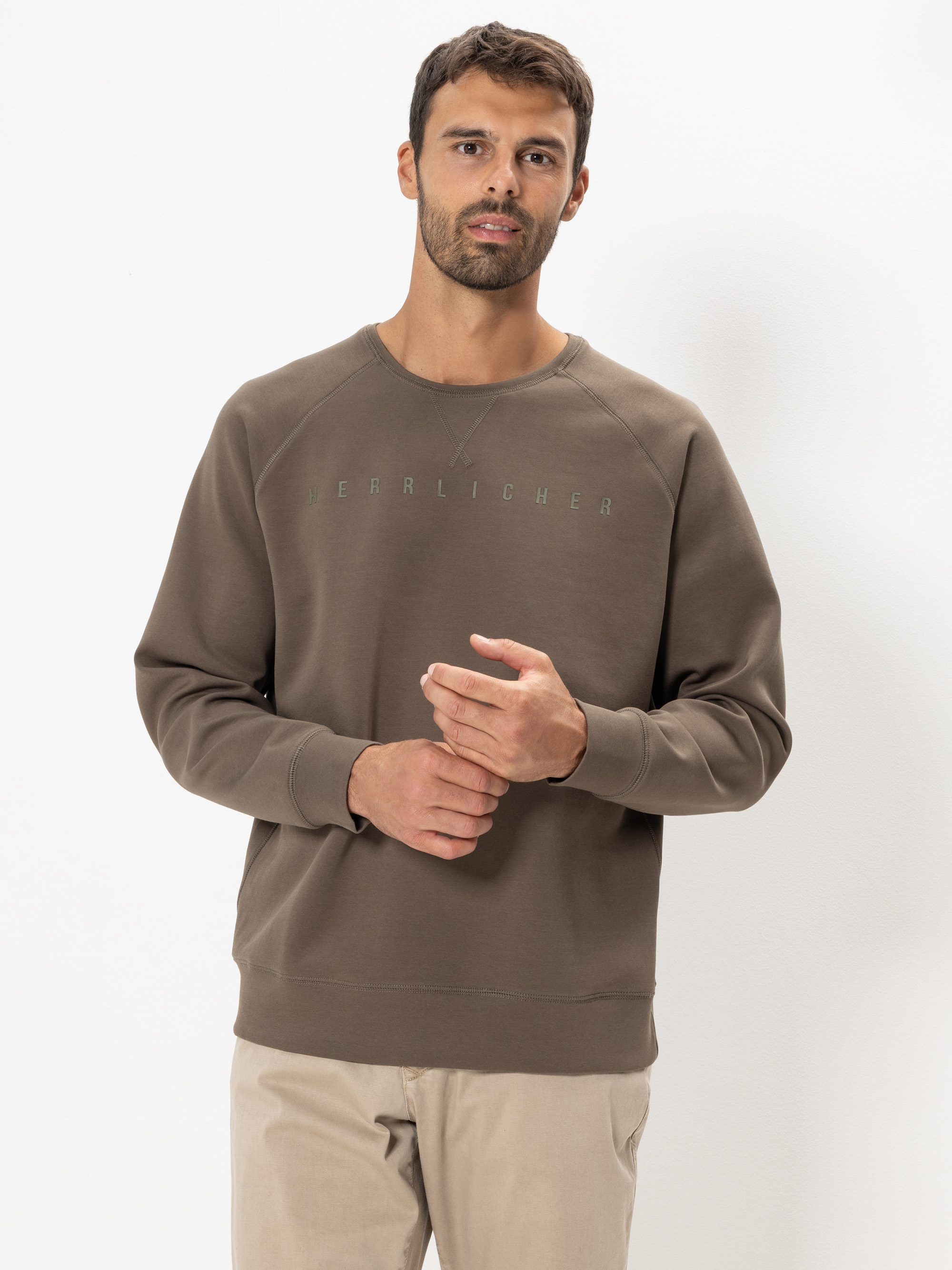 Ein Mann mit kurzen braunen Haaren und einem Bart trägt ein braunes Sweatshirt mit der Aufschrift HERBLICHER und eine beigefarbene Hose, die vor einem schlichten weißen Hintergrund steht.