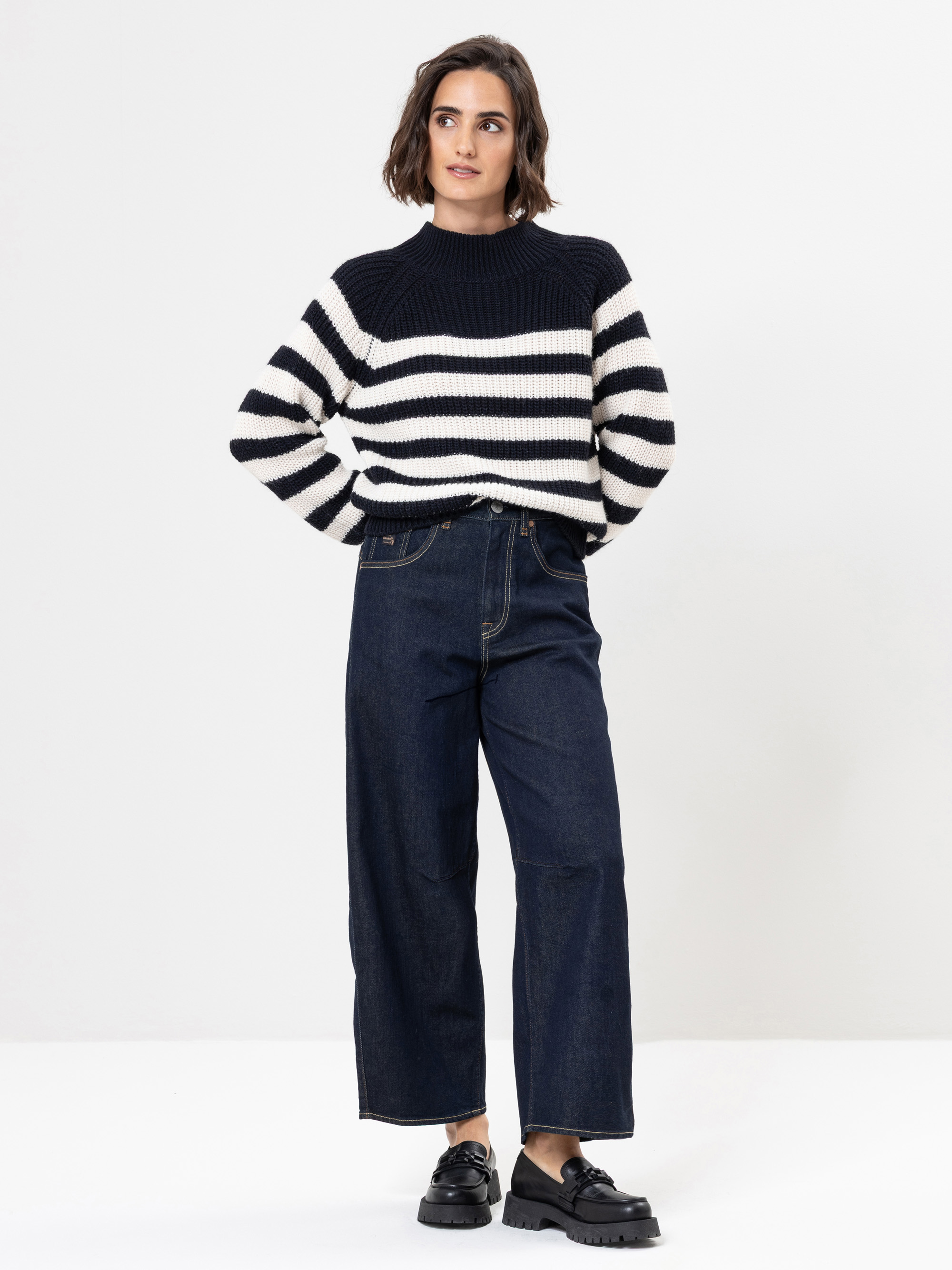 Eine Frau steht vor einem weißen Hintergrund und trägt einen schwarz-weiß gestreiften Pullover, dunkelblaue Jeans mit weitem Bein und schwarze Schuhe. Sie hat kurzes braunes Haar und ihre Hände sind in den Taschen verstaut.
