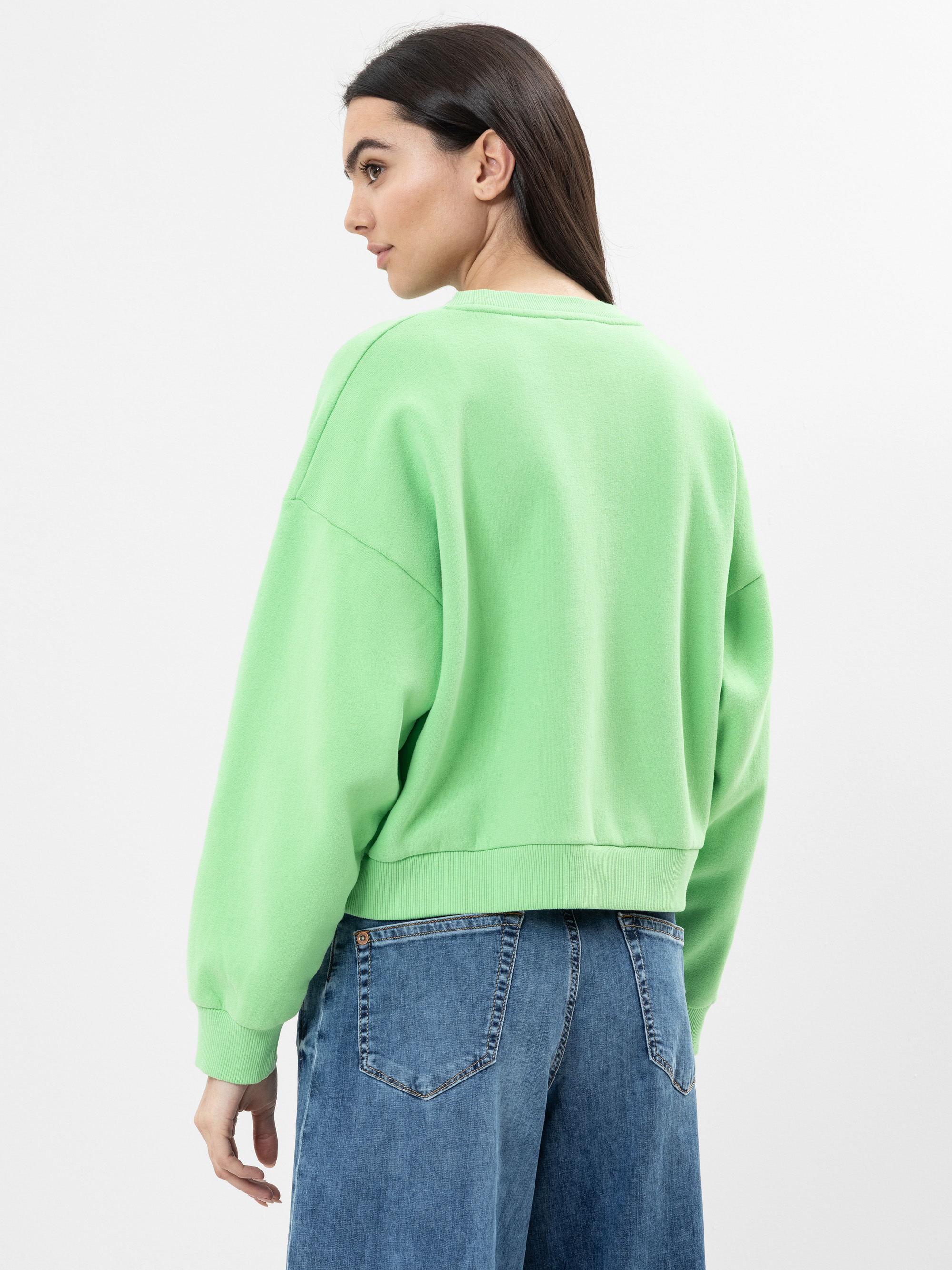 Eine Frau mit langen braunen Haaren steht leicht abgewandt in einem lockeren hellgrünen Sweatshirt und einer hoch sitzenden blauen Jeans vor einem schlichten weißen Hintergrund.