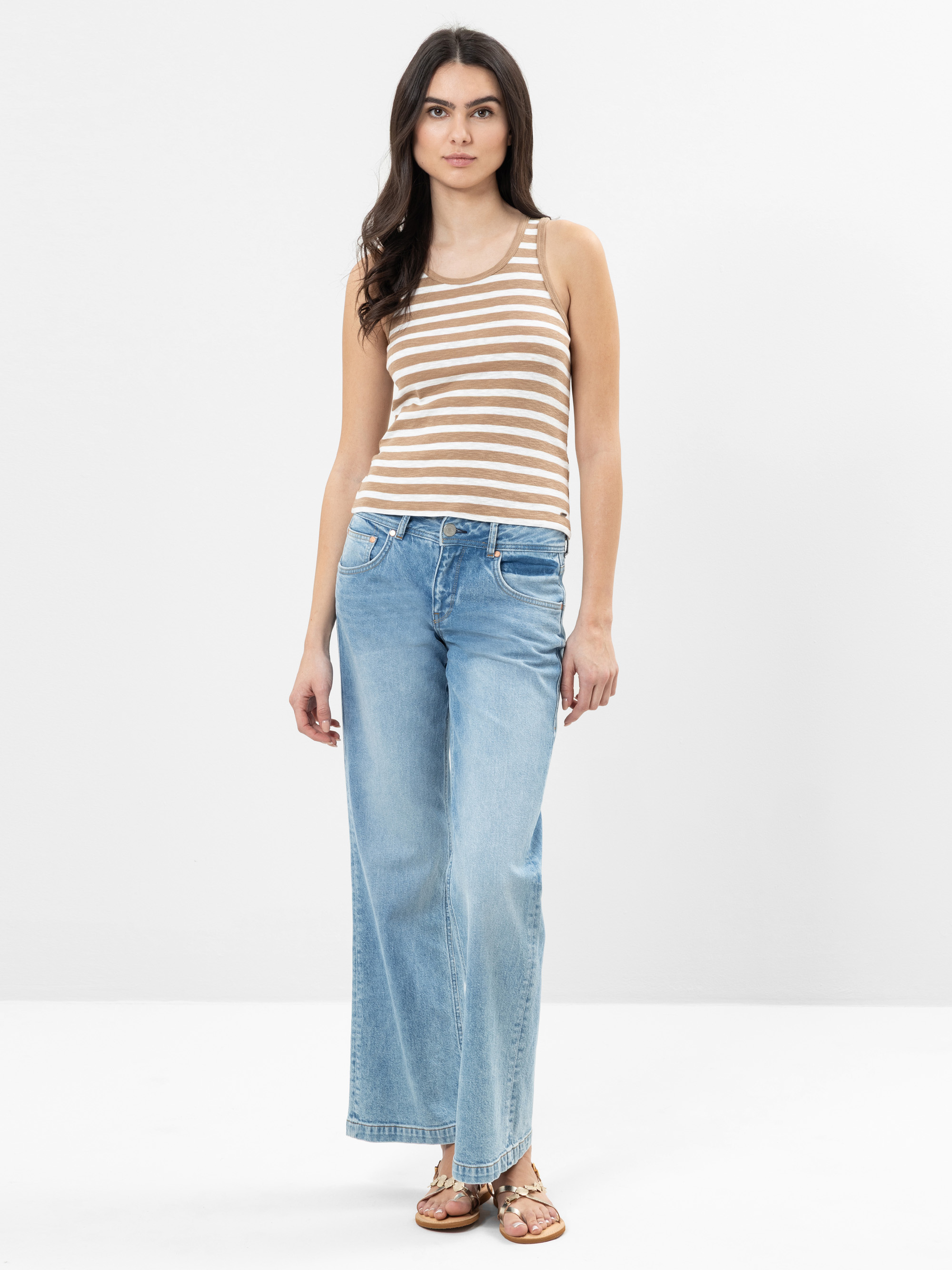 Eine Frau steht vor einem schlichten weißen Hintergrund und trägt ein beige-weiß gestreiftes ärmelloses Oberteil, hellblaue Jeans mit weitem Bein und braune Sandalen. Sie hat langes dunkles Haar und einen neutralen Gesichtsausdruck.