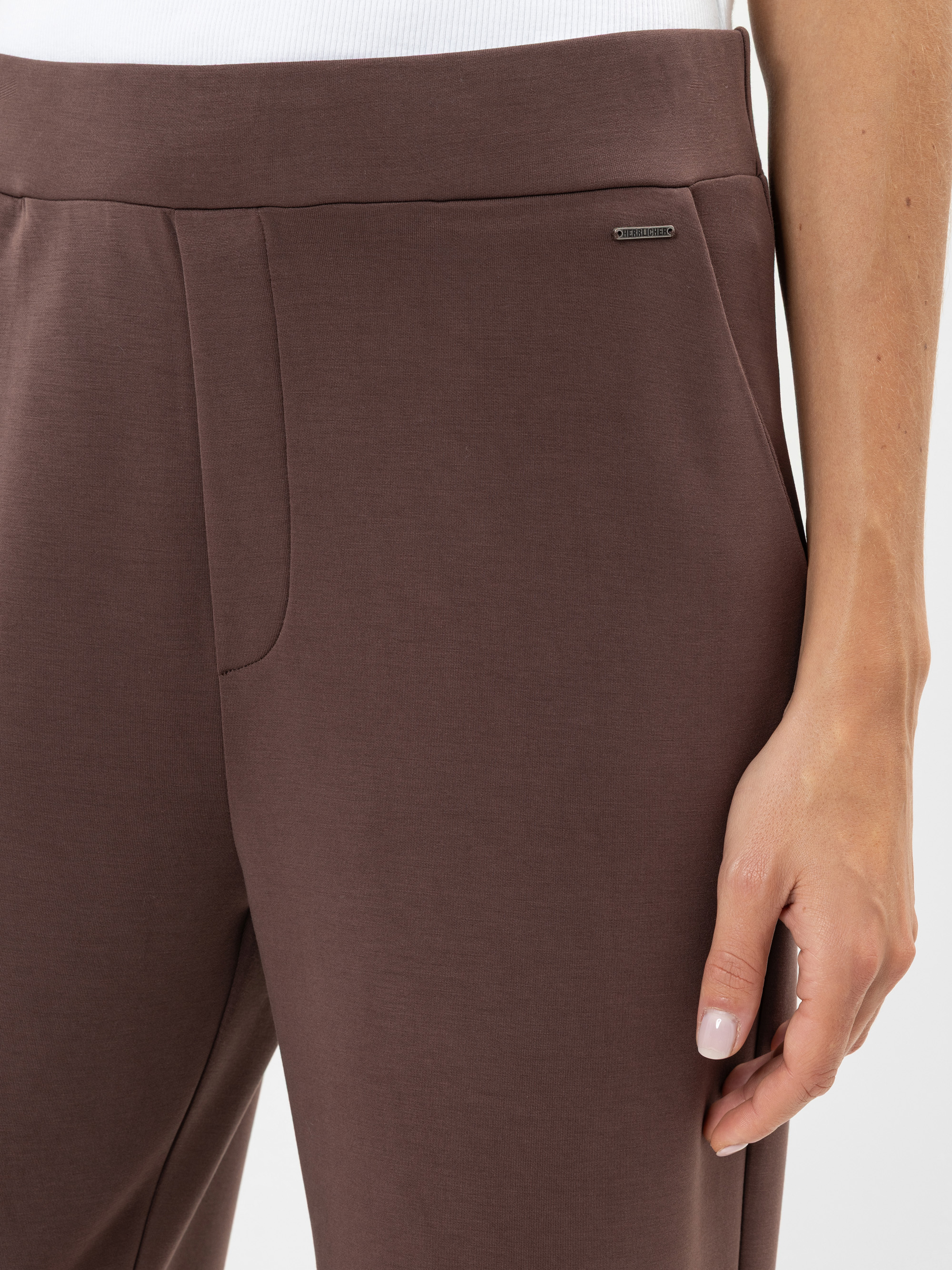 Nahaufnahme einer Person, die eine braune Hose und ein weißes Oberteil trägt und deren rechte Hand auf ihrem Oberschenkel ruht. In der Nähe der Tasche ist ein kleines metallisches Markenzeichen zu sehen.