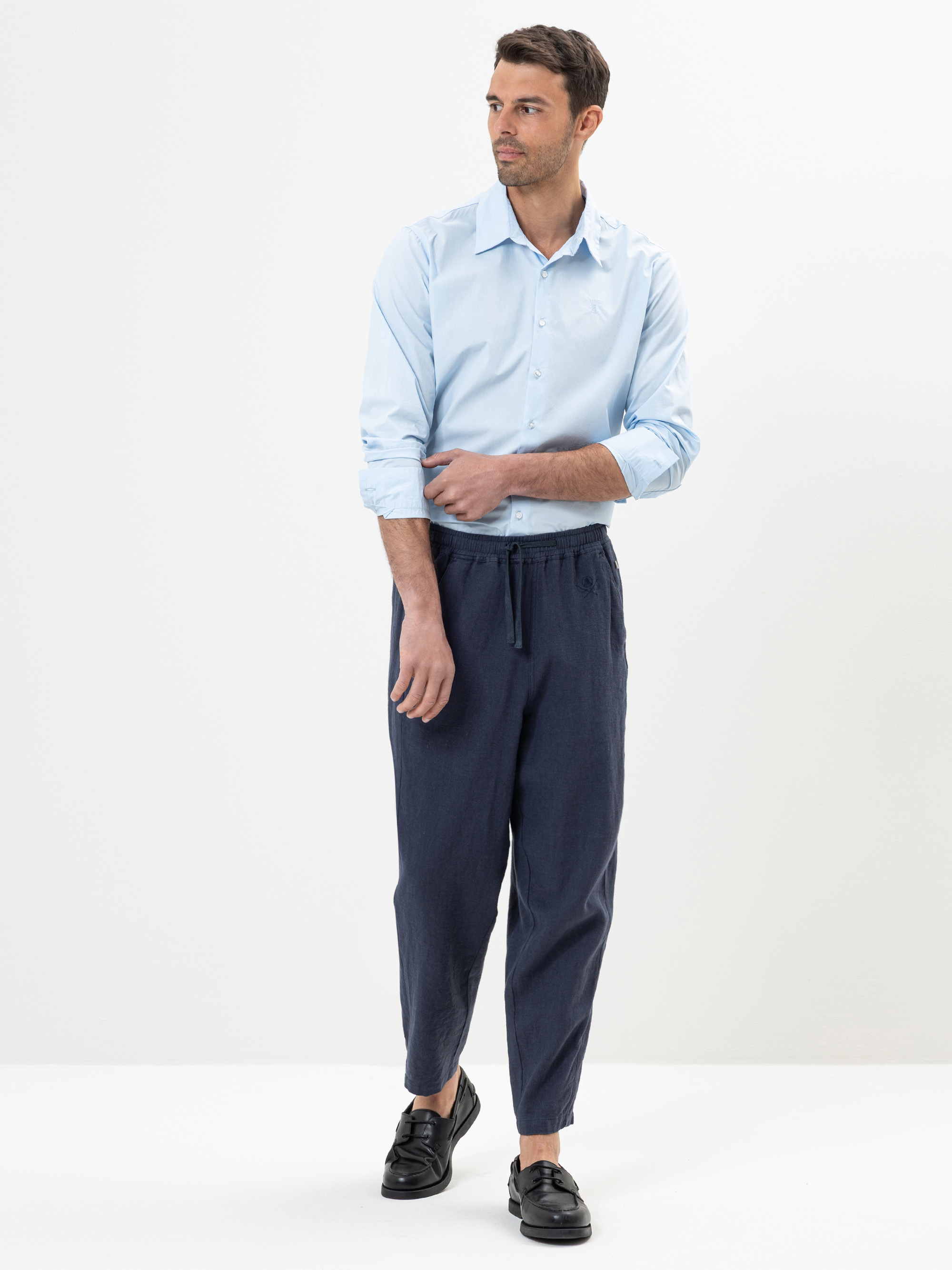 Ein Mann, der ein hellblaues Hemd mit gekrempelten Ärmeln, eine dunkelblaue Hose mit Kordelzug und schwarze Slipper trägt, steht vor einem schlichten weißen Hintergrund und schaut leicht nach links.