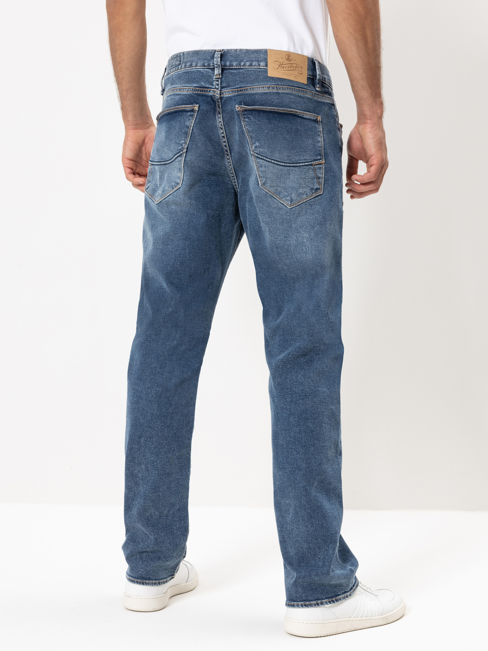 Ein Mann in hellblauen Jeans mit geradem Bein, weißem T-Shirt und weißen Turnschuhen, von hinten vor einem schlichten weißen Hintergrund dargestellt.