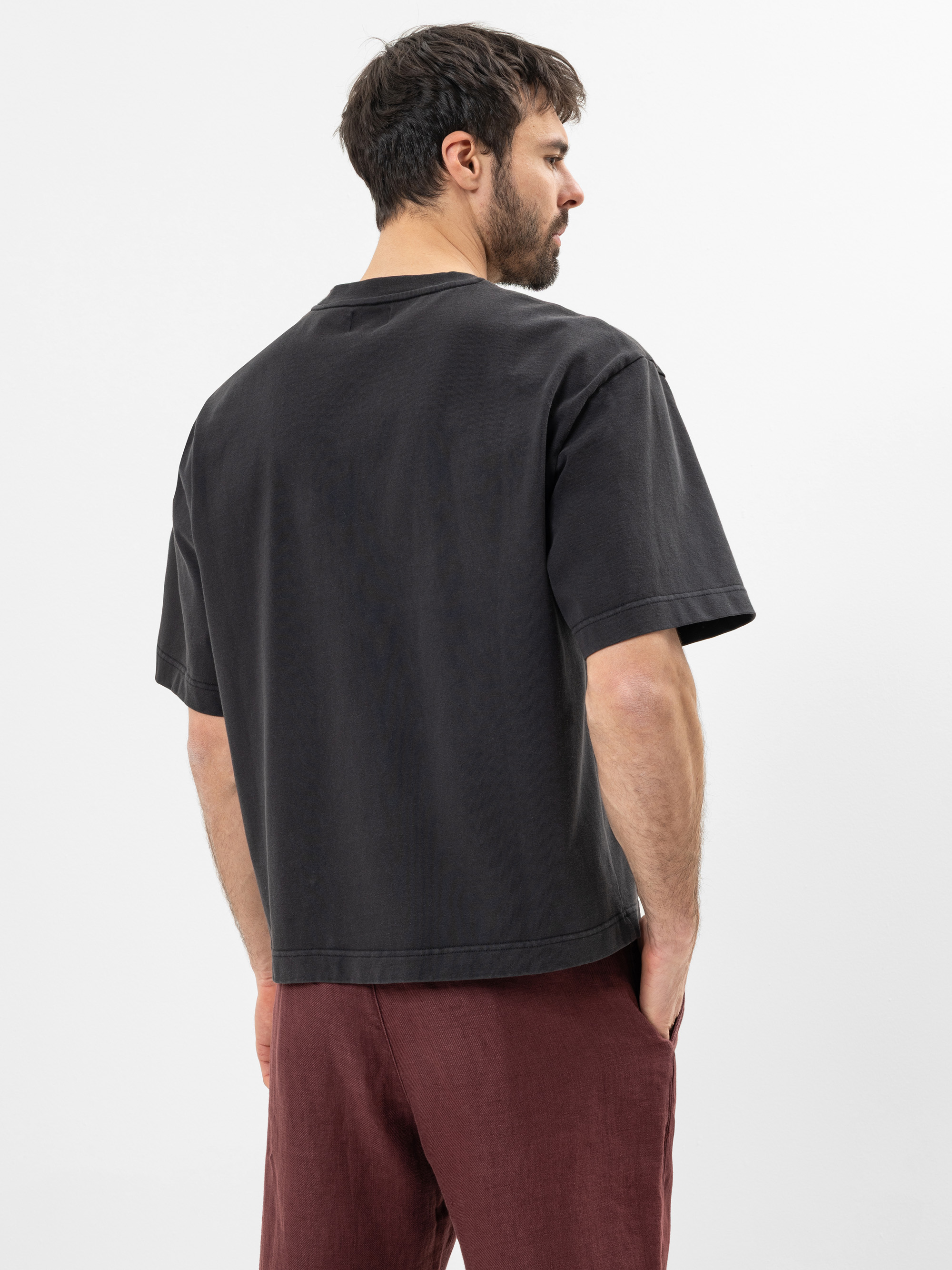 Ein Mann mit dunklem Haar und Bart steht mit dem Rücken zur Kamera, trägt ein locker sitzendes schwarzes T-Shirt und eine kastanienbraune Hose, die Hände in den Taschen, vor einem schlichten weißen Hintergrund.