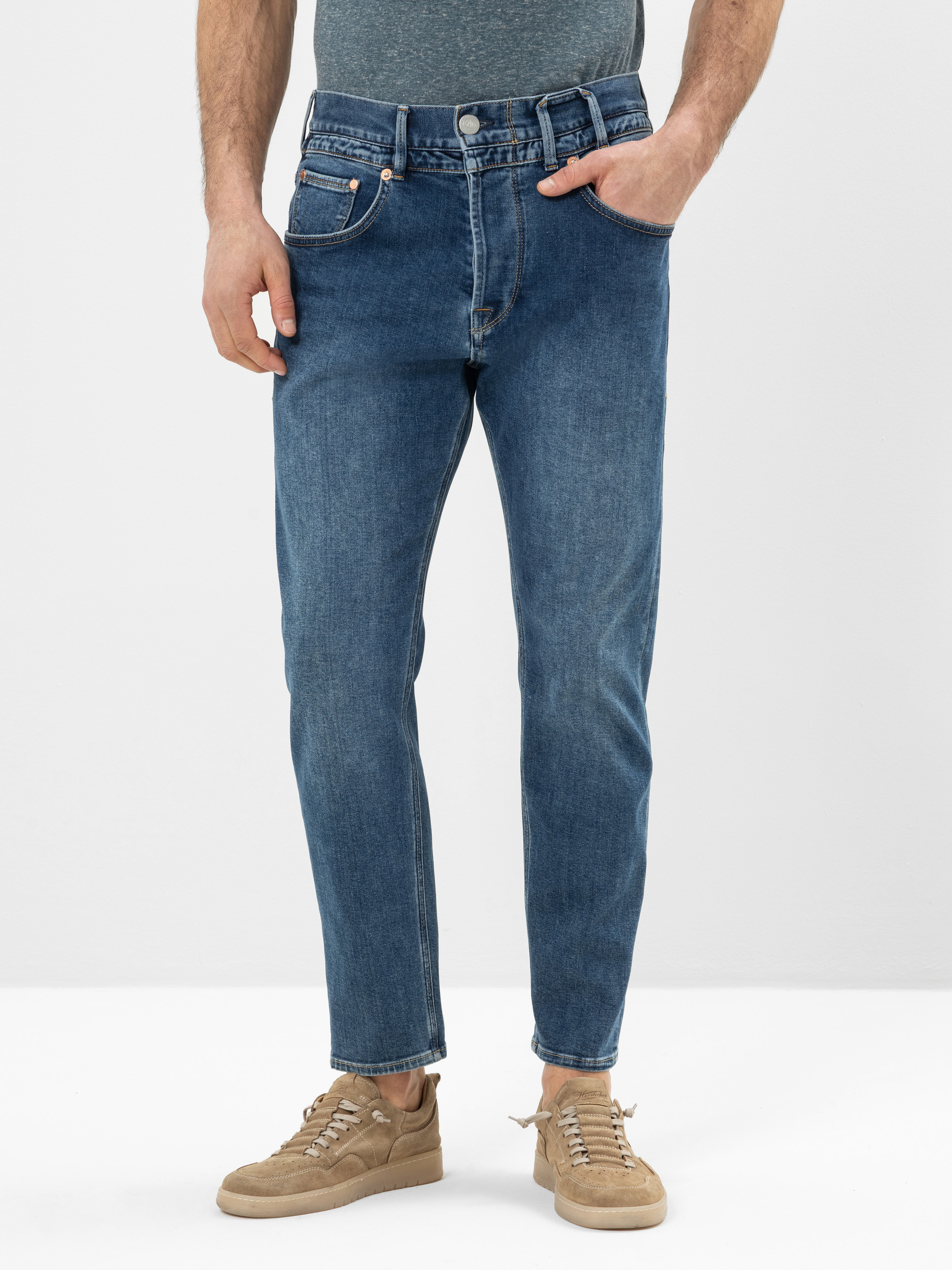 Eine Person, die blaue Jeans, ein graues Hemd und hellbraune Turnschuhe trägt, steht mit einer Hand in der Tasche vor einem schlichten hellen Hintergrund. Das Foto ist von der Brust abwärts beschnitten.