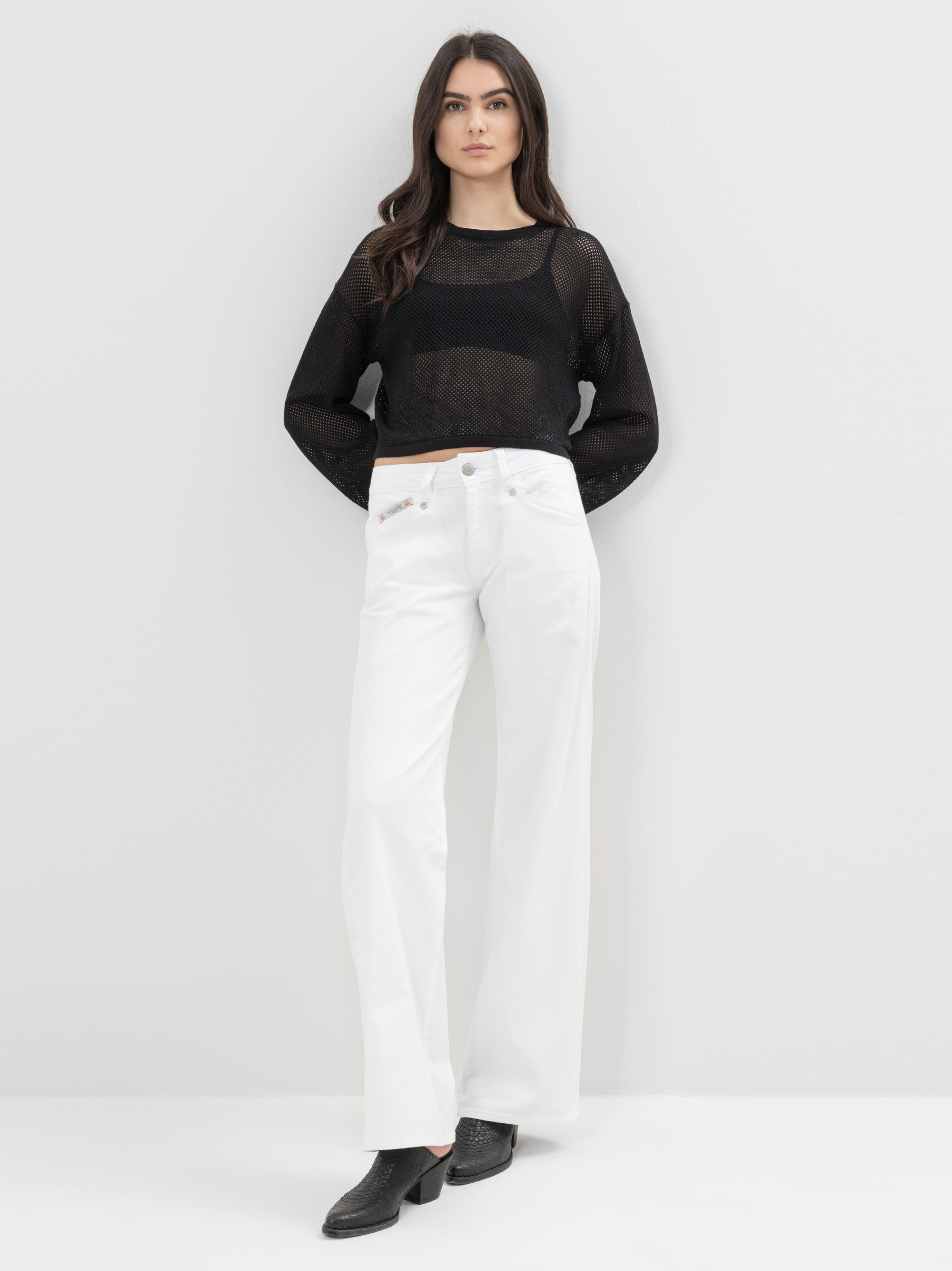 Eine Frau steht vor einer schlichten weißen Wand. Sie trägt ein durchsichtiges schwarzes, langärmeliges Oberteil, eine weiße, hochgeschlossene Hose mit weitem Bein und schwarze Schuhe. Sie blickt mit den Händen in den Taschen nach vorne.