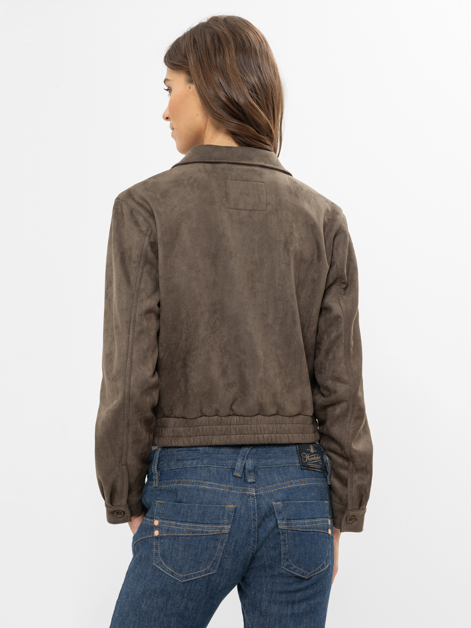 Eine Frau mit langen braunen Haaren, von hinten gesehen, trägt eine braune Wildlederjacke und blaue Jeans vor einem schlichten weißen Hintergrund.