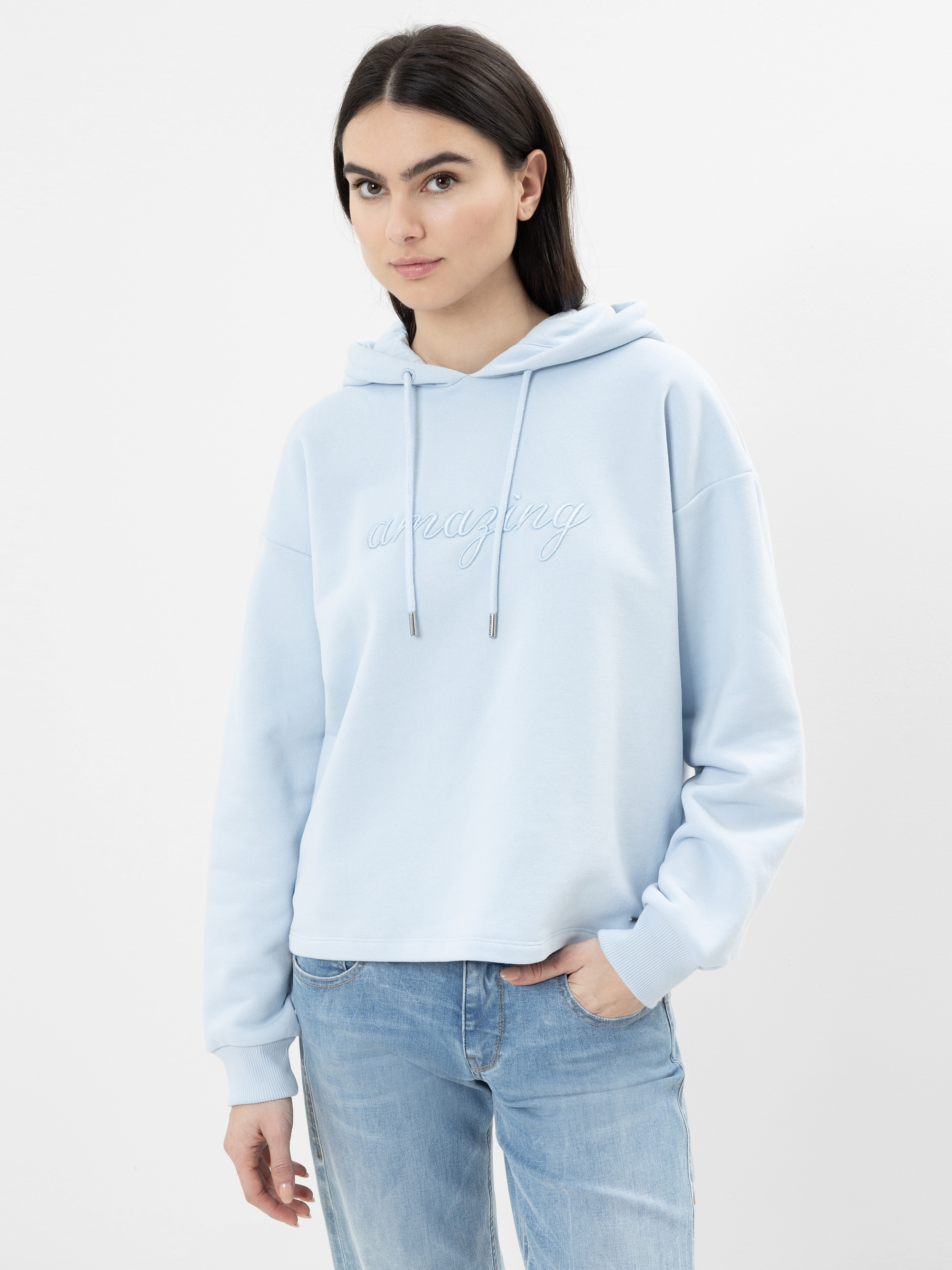 Eine Frau mit langen dunklen Haaren, die einen hellblauen Kapuzenpullover mit der Aufschrift "amazing" und hellblaue Jeans trägt, steht vor einem schlichten weißen Hintergrund und schaut selbstbewusst in die Kamera.