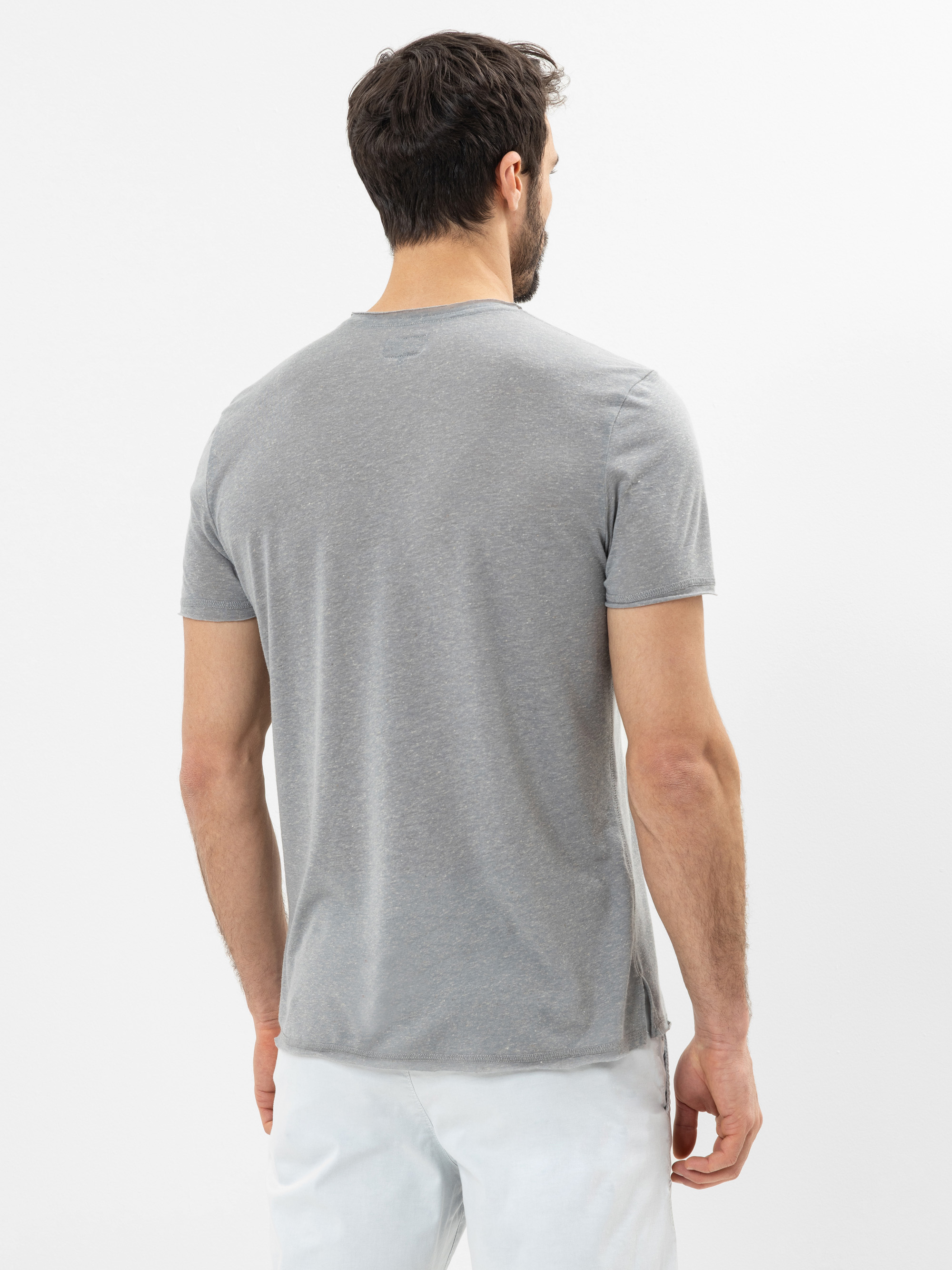 Ein Mann mit kurzen dunklen Haaren wird von hinten gezeigt. Er trägt ein schlichtes hellgraues, kurzärmeliges T-Shirt und eine weiße Hose und steht vor einem weißen Hintergrund.