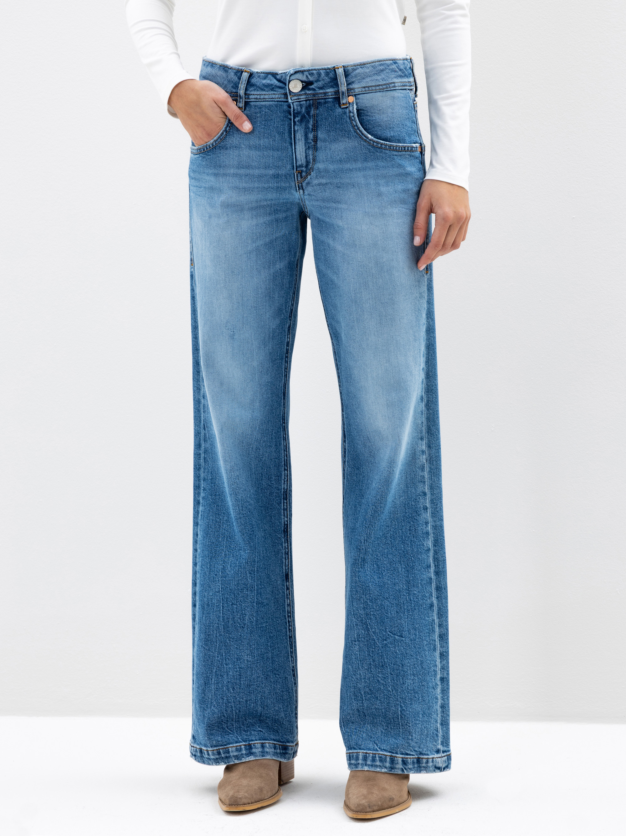 Eine Person, die ein weißes langärmeliges Oberteil, hellblaue Jeans mit weitem Bein und hellbraune Schuhe trägt, steht vor einem schlichten weißen Hintergrund und hat eine Hand in der Tasche.