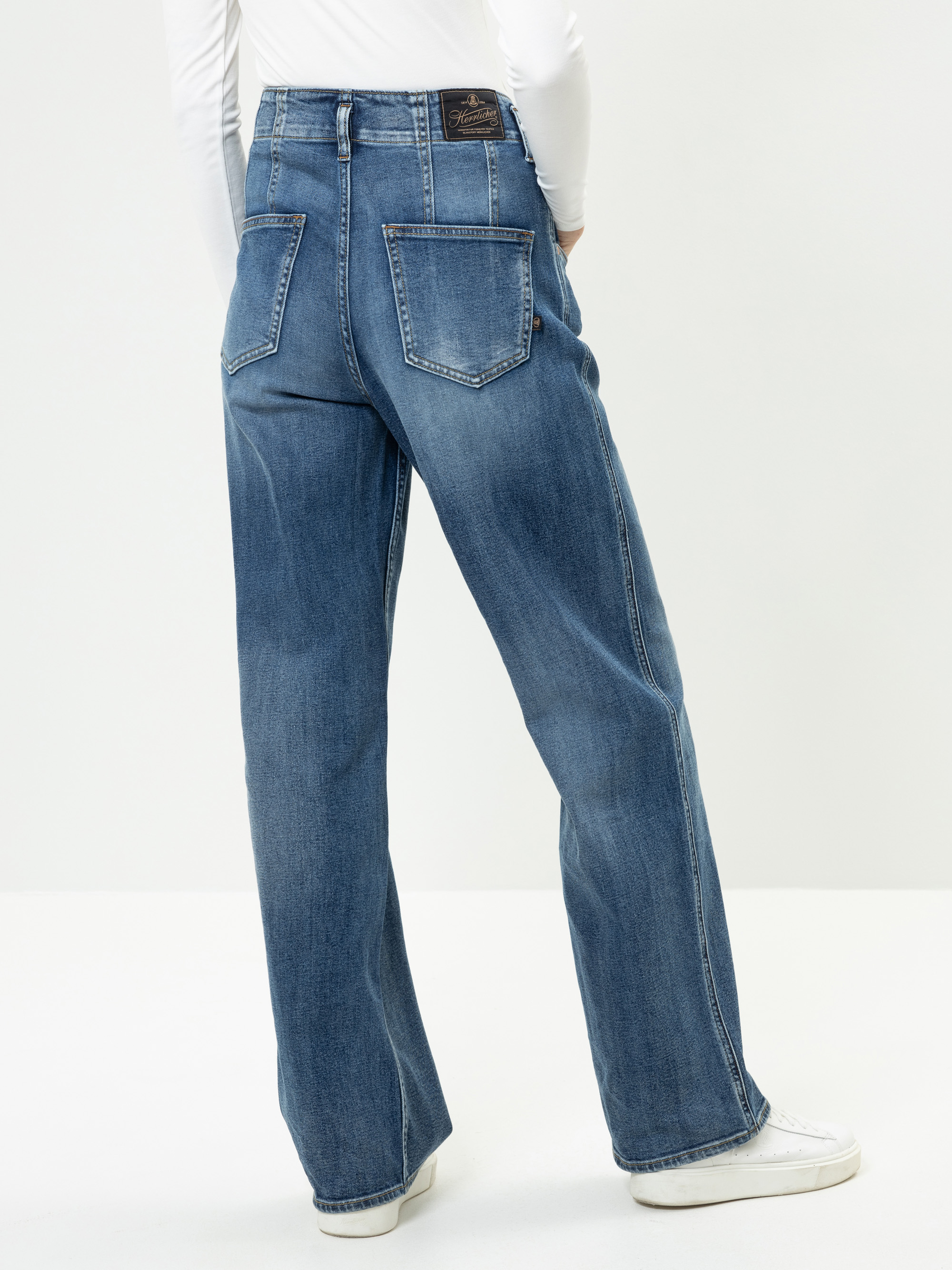 Eine Person, die eine blaue Jeans mit hoher Taille und weitem Bein sowie weiße Turnschuhe trägt, steht mit dem Rücken zur Kamera vor einem schlichten hellen Hintergrund. Die Jeans hat große Gesäßtaschen und einen Markenaufnäher am Bund.