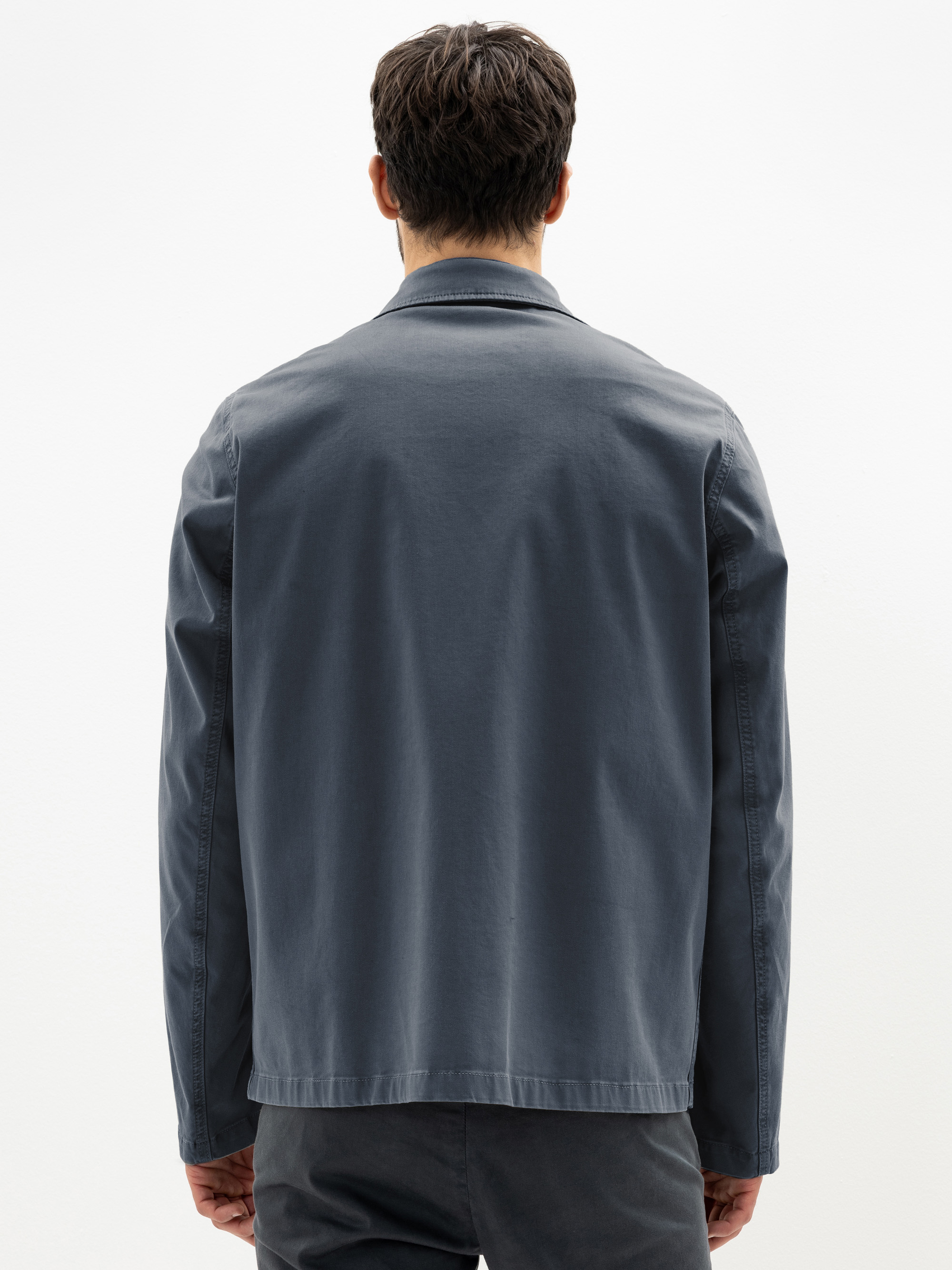 Ein Mann mit kurzen braunen Haaren steht mit dem Rücken zur Kamera, trägt eine dunkelgraue Jacke und eine dazu passende dunkle Hose vor einem schlichten weißen Hintergrund.