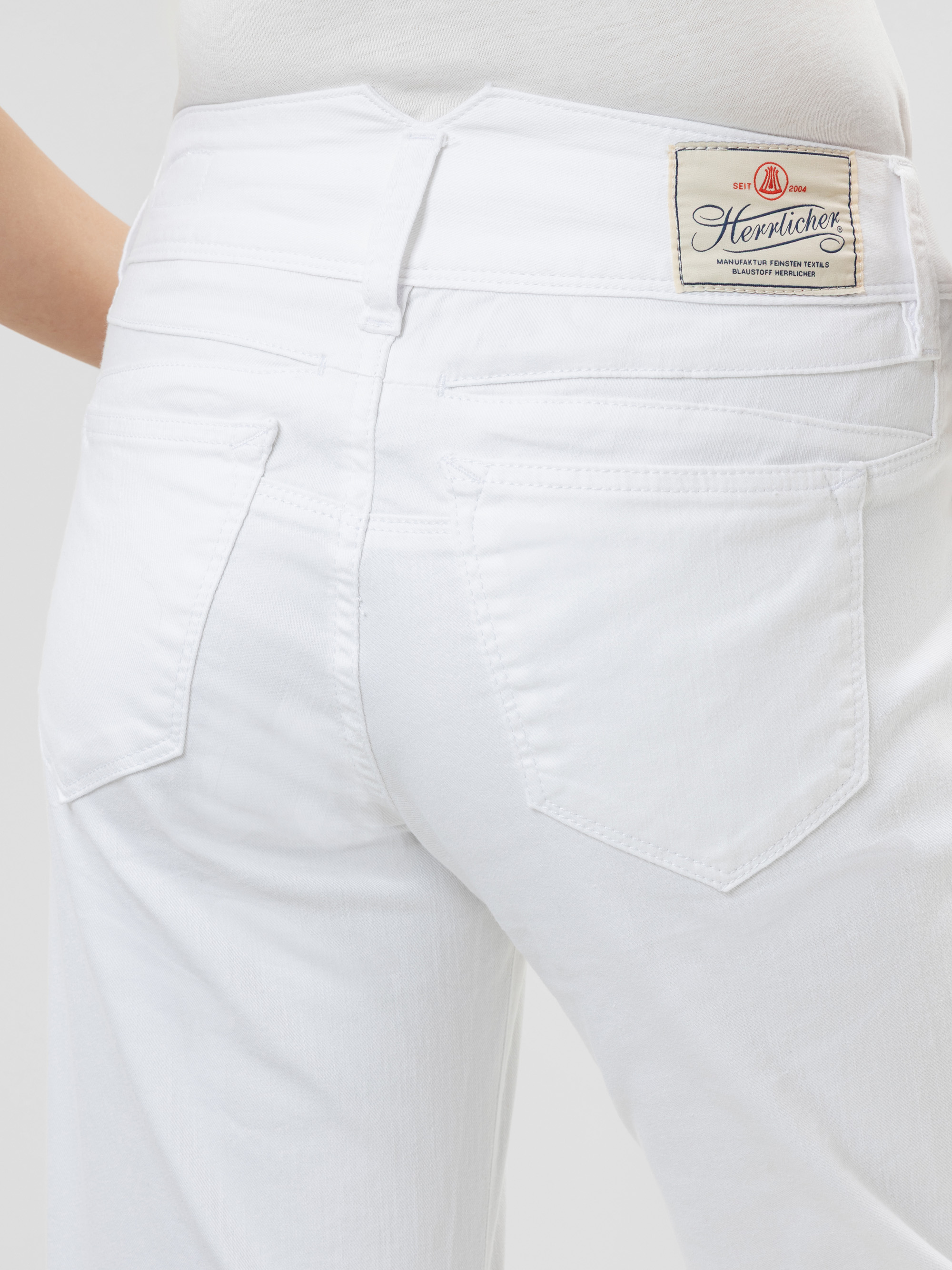 Nahaufnahme einer Person, die eine weiße Jeans und ein weißes Oberteil trägt, mit Blick auf den Rücken. Die Jeans hat zwei Gesäßtaschen und ein beigefarbenes Etikett am Bund mit dem Markennamen Herrlicher.