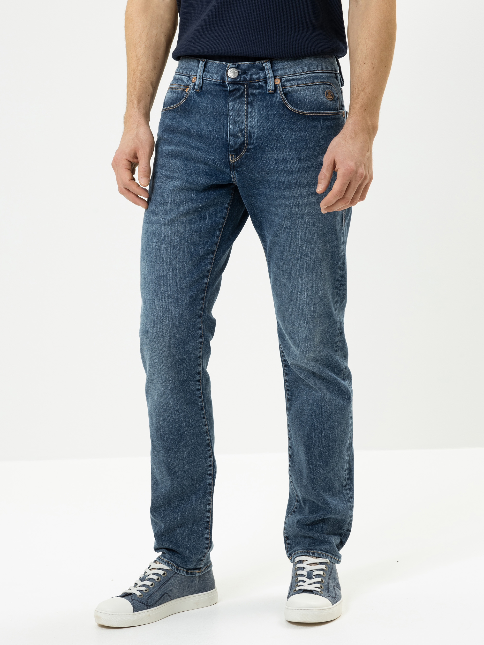 Ein Mann, der eine blaue Jeans mit geradem Bein, ein dunkles Hemd und grau-weiße Turnschuhe trägt, steht vor einem schlichten weißen Hintergrund. Seine Hände liegen entspannt an den Seiten, und nur sein Körper ist von den Schultern abwärts zu sehen.