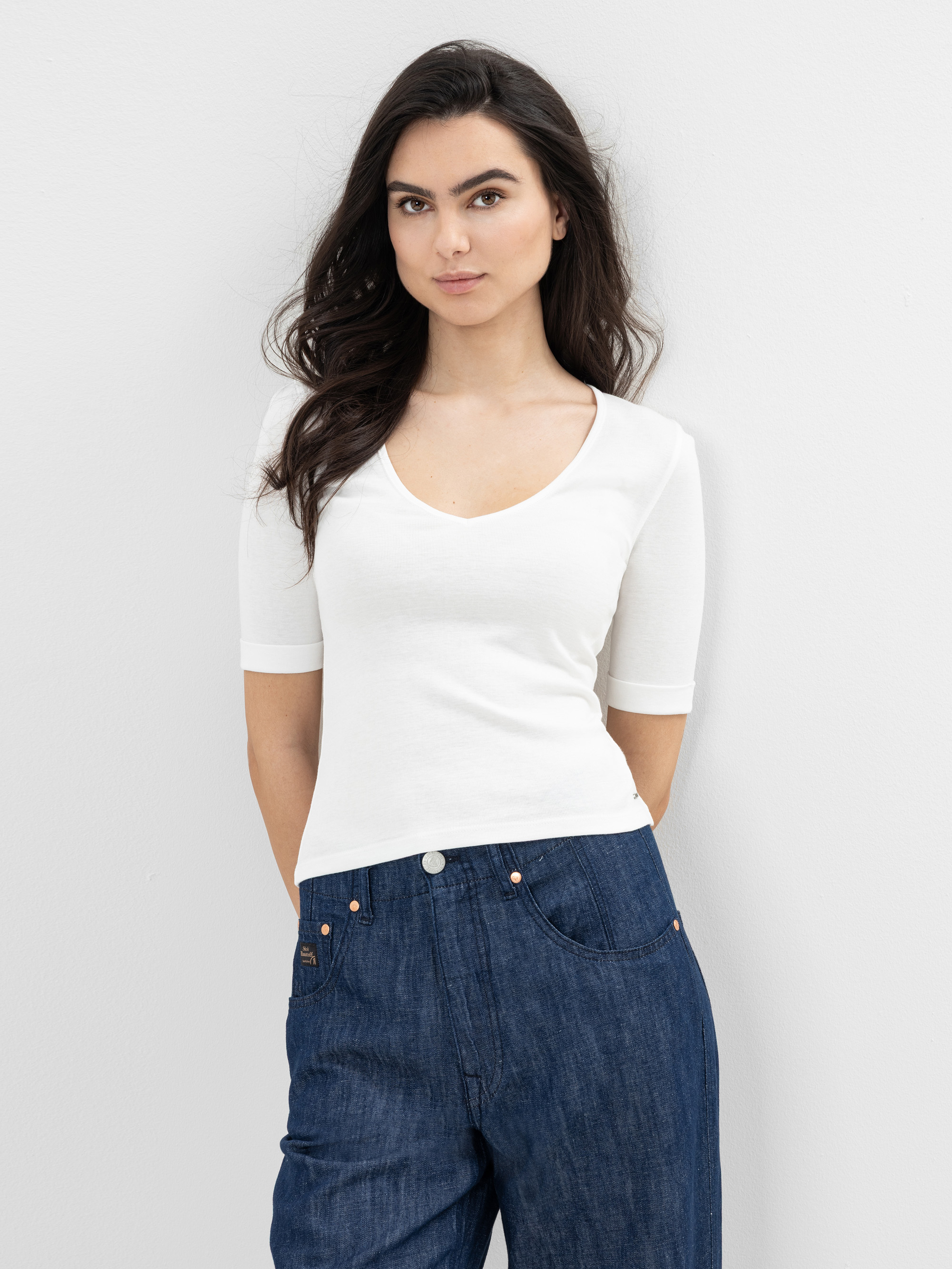 Eine Frau mit langen dunklen Haaren, die ein weißes, kurzärmeliges Oberteil und blaue Jeans trägt, steht vor einer schlichten hellen Wand und blickt selbstbewusst in die Kamera.