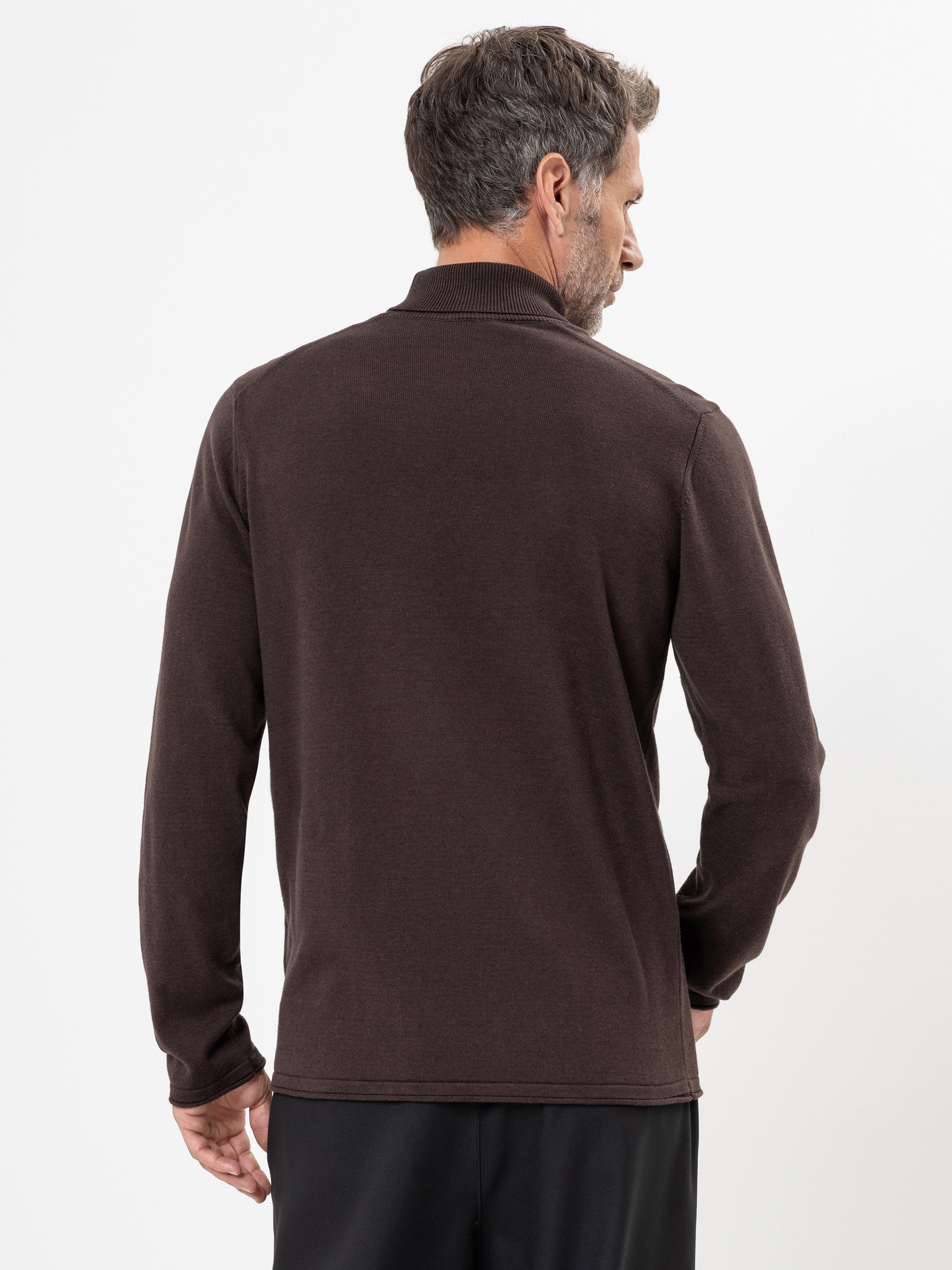 Ein Mann mit kurzen Haaren steht mit dem Rücken zur Kamera, trägt ein langärmeliges dunkelbraunes Polohemd und eine schwarze Hose vor einem schlichten weißen Hintergrund.