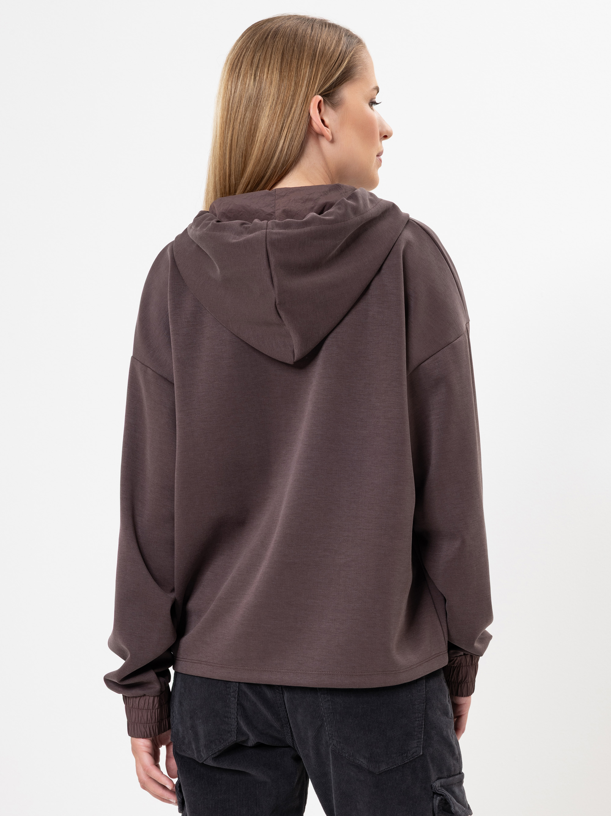 Eine Frau mit glattem, hellbraunem Haar wird von hinten gezeigt. Sie trägt ein locker sitzendes, dunkelbraunes Sweatshirt mit Kapuze und eine schwarze Hose vor einem schlichten weißen Hintergrund.