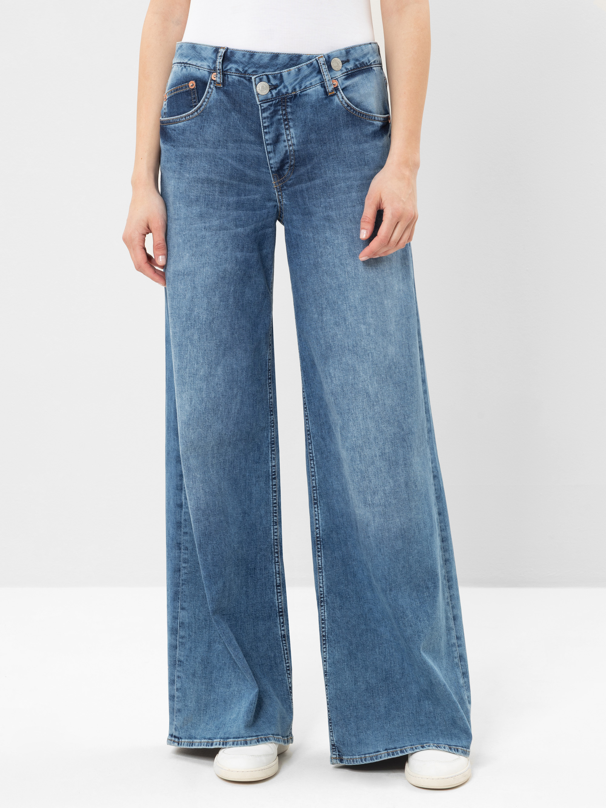 Eine Person, die eine blaue Jeans mit hoher Taille und weitem Bein sowie weiße Turnschuhe trägt, steht vor einem schlichten hellgrauen Hintergrund. Die obere Hälfte ihres Körpers ist nicht sichtbar.