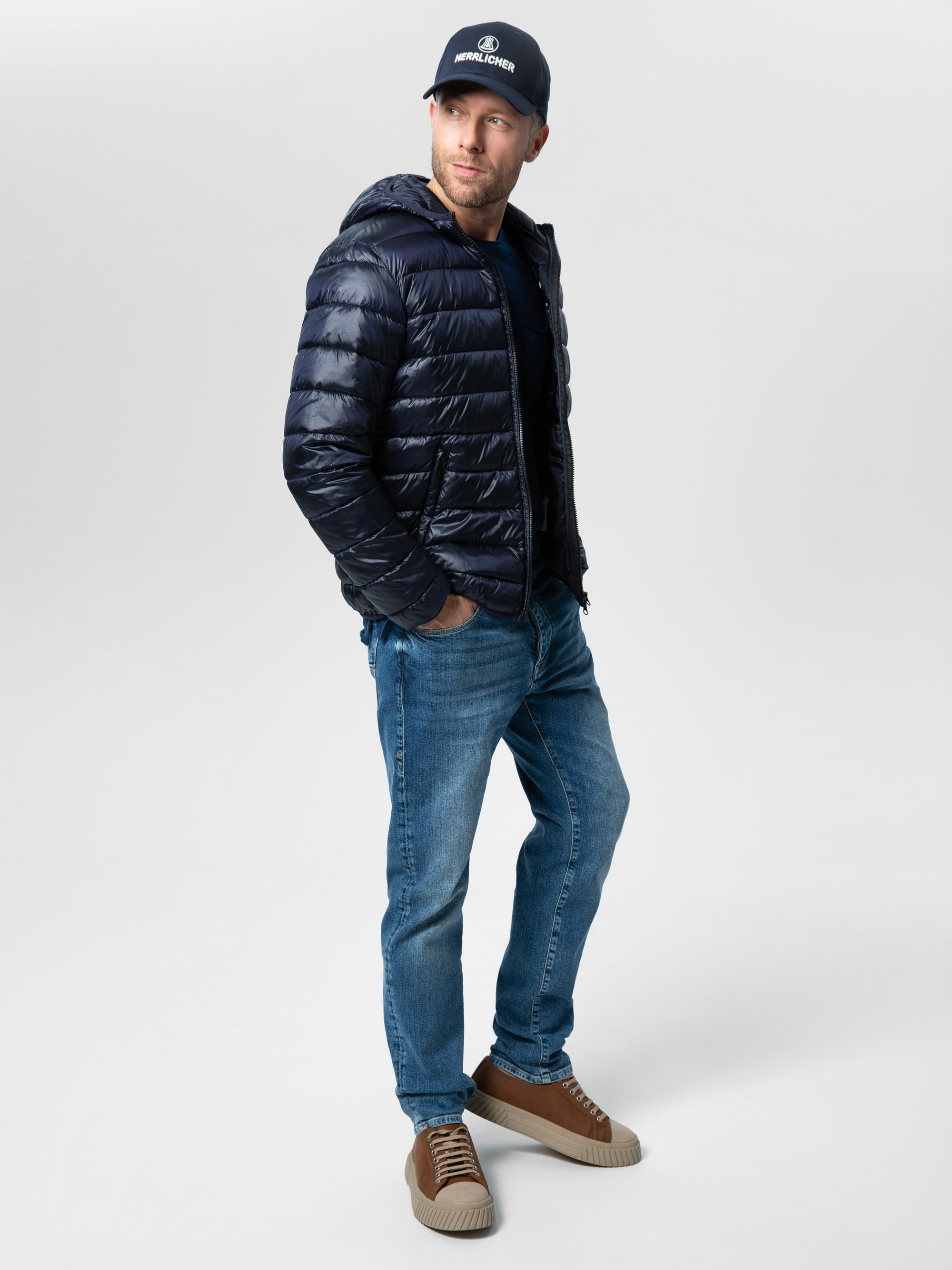 Ein Mann, der eine marineblaue Pufferjacke, blaue Jeans, hellbraune Turnschuhe und eine schwarze Kappe trägt, steht mit einer Hand in der Tasche und blickt zur Seite vor einem schlichten hellgrauen Hintergrund.