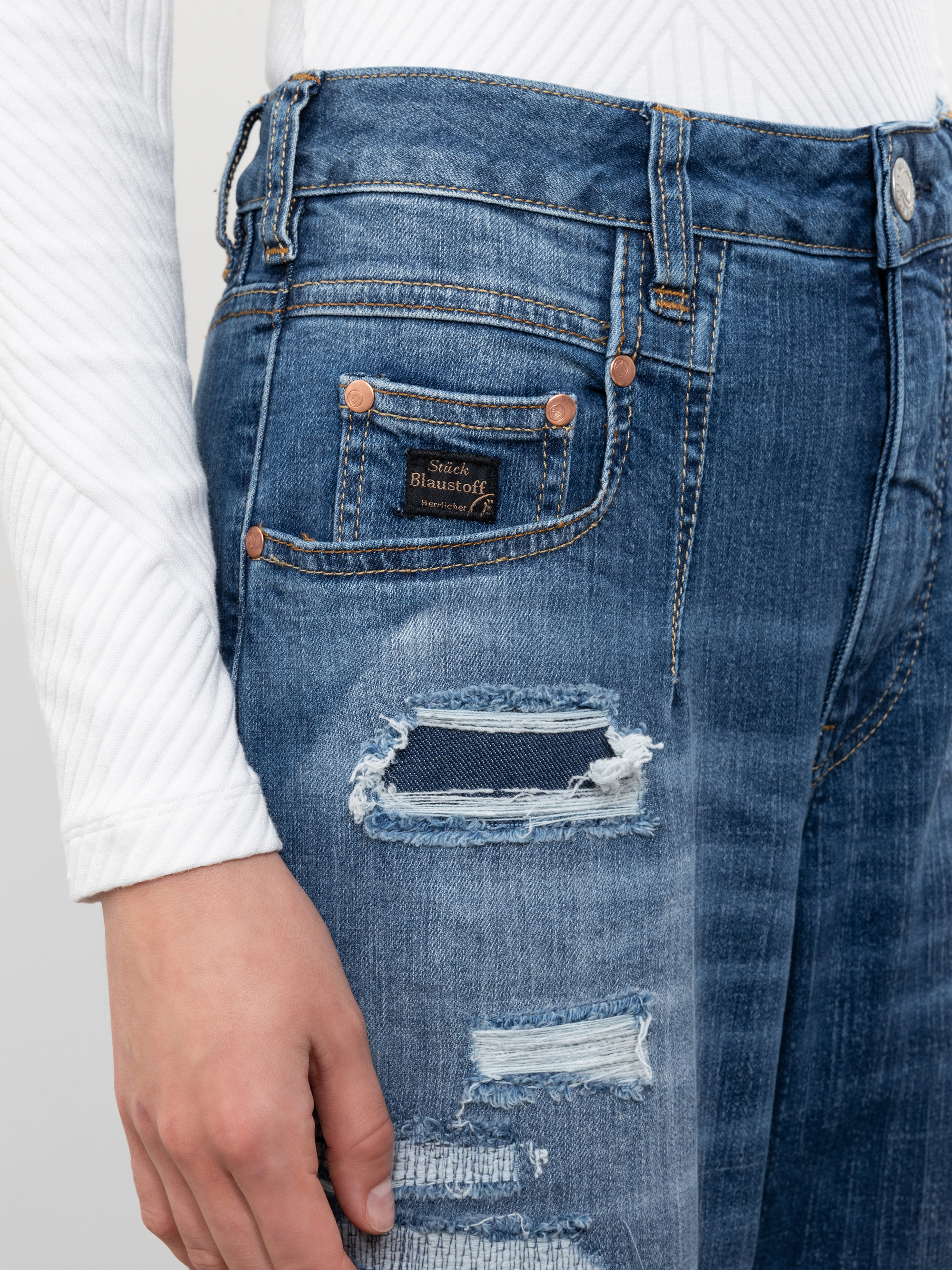 Eine Person, die ein weißes, geripptes, langärmeliges Hemd und eine blaue, abgenutzte Jeans mit mehreren Rissen und einem kleinen Aufnäher in der Nähe der Vordertasche trägt.