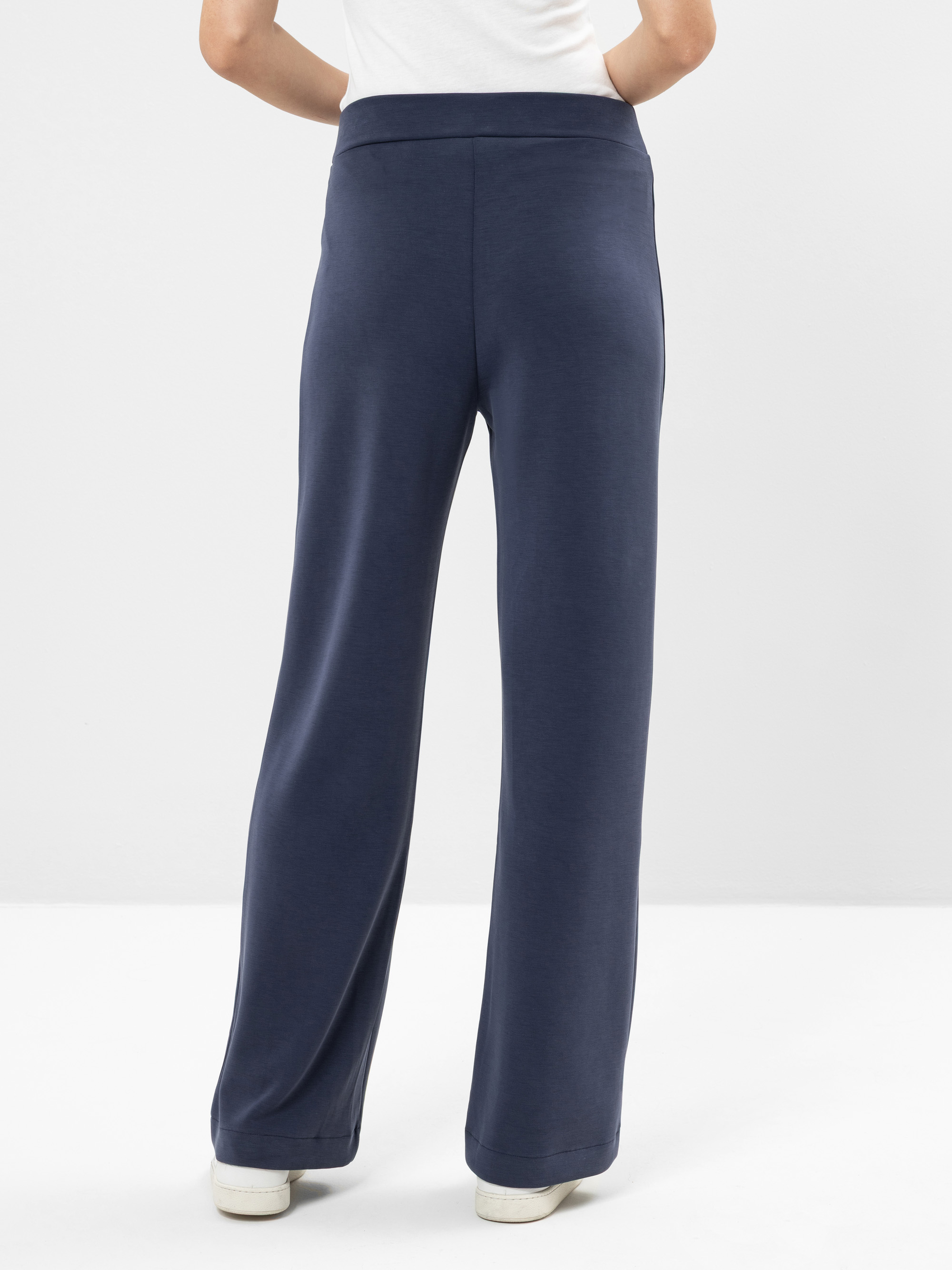 Eine Person, die eine marineblaue Hose mit weitem Bein und weiße Turnschuhe trägt, wird von hinten gezeigt und steht vor einem schlichten weißen Hintergrund.